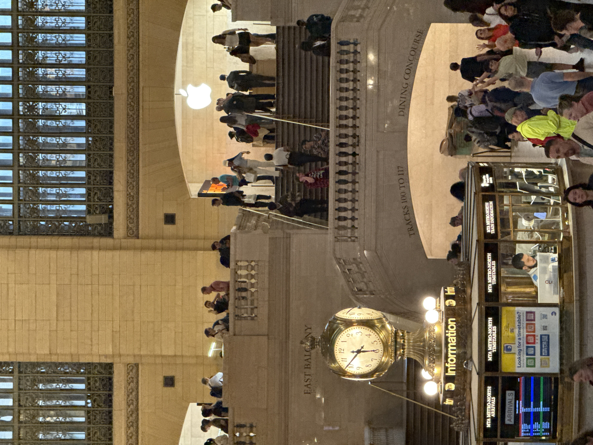 Here's a detailed description of the image:
**Scene:** The image shows the interior of Grand Central Terminal in New York City.
**People:** There are numerous people visible throughout the image. They are walking on staircases, looking at train information, and standing on a balcony.
**Objects:** Key objects in the scene include:
* A large, ornate clock in the foreground.
* An information booth with digital displays showing train arrival and departure times.
* Staircases leading to different levels within the terminal.
* Balconies overlooking the main concourse.
* Large windows with intricate metalwork.
* The Apple Store logo, mounted on the wall
**Environment:** The interior is grand and spacious, with high ceilings and stone walls. The light is warm, suggesting artificial lighting. The overall atmosphere is busy and bustling, indicative of a transportation hub.
**Interactions:** People are interacting with the environment by walking around and presumably making their way to different destinations. Some people are likely looking at the information screens to check train schedules.