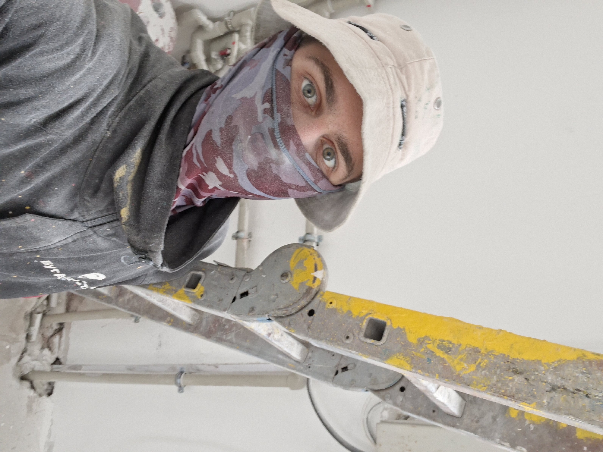 A close-up, upward-angled shot shows a person wearing a gray hooded sweatshirt with paint splatters, a camouflage-patterned face covering, and a light-colored hat. The person is standing on a yellow and gray ladder. Background shows white walls with pipes and plaster. Location and city are unknown.