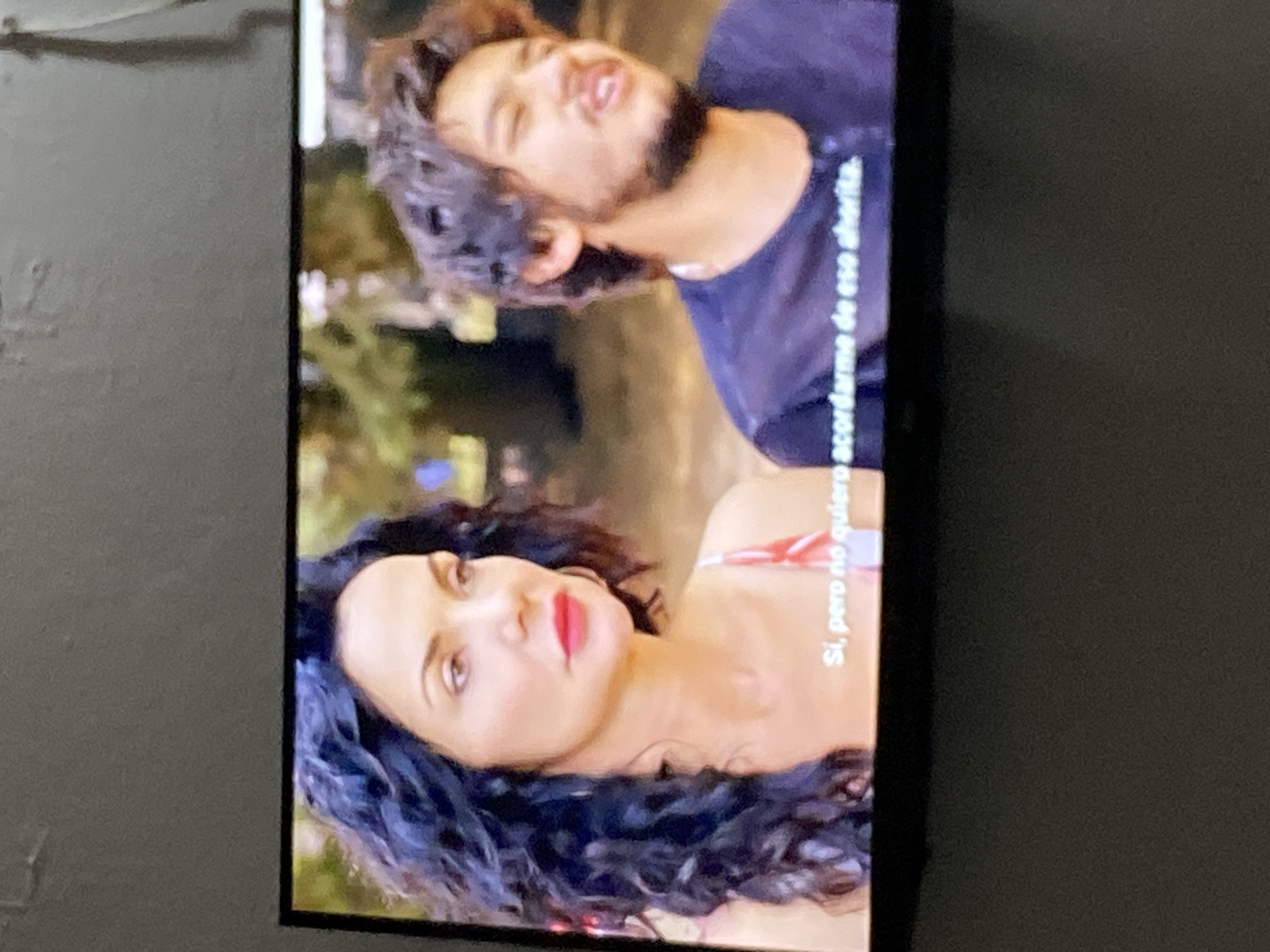 This media file shows a close-up shot of two people, a man and a woman, on what appears to be a television screen. The image is oriented vertically, with the subjects appearing sideways.
The man, in the upper portion of the image, has dark, curly hair and a beard. He is wearing a dark t-shirt and his head is tilted back, with his mouth open as if speaking or laughing. His expression appears lively.
Below him, the woman has long, dark, curly hair and is wearing makeup, including bright red lipstick. She is looking upwards and slightly to the side, with a thoughtful or perhaps slightly pensive expression. She is wearing a strappy garment with a red and white pattern.
There is visible text on the right side of the image, oriented vertically. It appears to be in Spanish and reads: "Sí, pero no quiero acordarme de eso ahorita." This translates to "Yes, but I don't want to remember that right now."
The background is blurred, suggesting an outdoor setting with some greenery and possibly artificial lights, hinting at an evening or nighttime scene. The lighting on the subjects is soft. The overall mood is intimate, possibly suggesting a conversation or a moment between the two individuals.