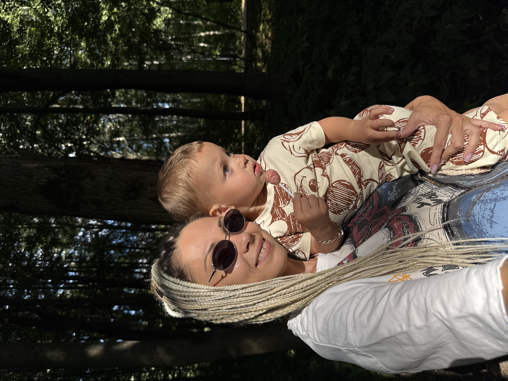 In the city of Rusanovo, Russia, a woman with blonde braided hair and sunglasses holds a young child while walking in a forest. The woman wears a white t-shirt over a graphic tee and a silver bracelet. The child is dressed in a light-colored onesie with a brown graphic print and holds a lollipop. The background consists of tall trees and dense foliage, with sunlight filtering through the canopy.