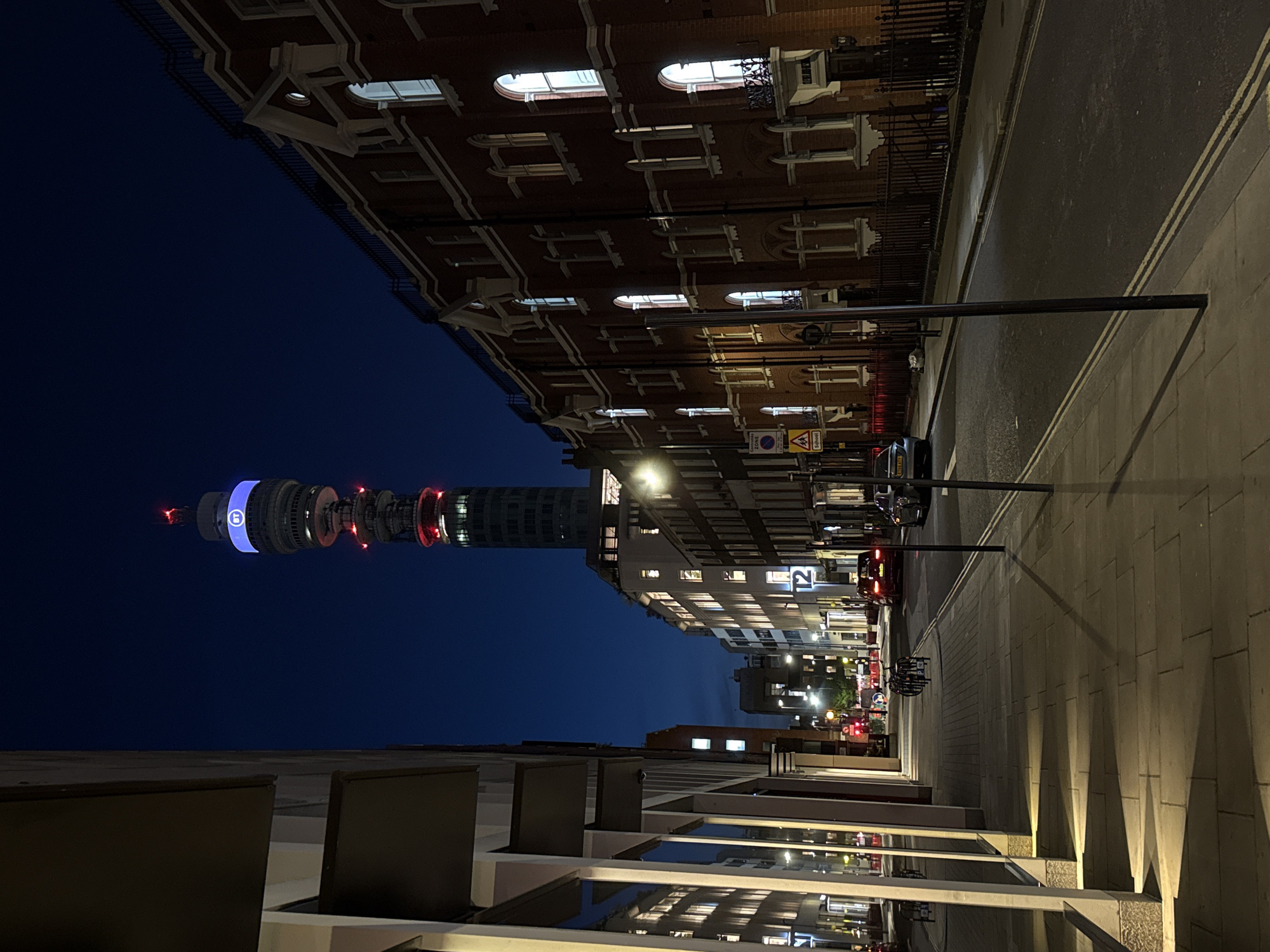 An urban night scene in London is captured, with the iconic BT Tower prominently featured against a dark indigo sky. The image appears to be rotated 90 degrees clockwise; when reoriented, the BT Tower stands tall on the left, its upper section illuminated by a distinctive blue band displaying the "BT" logo and accented with red lights.
To the right, a traditional multi-story brick building with ornate white window frames and balconies lines the street. Many of its windows are brightly lit, indicating interior activity. Further down the street, more modern buildings with glowing windows contribute to the cityscape.
In the foreground, a paved sidewalk on the right features a striking pattern of triangular shadows cast by wall-mounted uplights, creating a geometric visual effect. The street, made of dark asphalt, has visible double yellow lines and is partially illuminated by streetlights. Several vehicles, including parked cars with illuminated tail lights, are visible along the roadside.
Visible street signs include a yellow diamond "SCHOOL" warning sign, and a blue and white sign indicating "CONGESTION SIGNAL" with an "AA" logo. The time of day is clearly night or late evening, evidenced by the dark sky and pervasive artificial lighting. The clear sky offers no immediate indication of specific weather conditions, beyond a lack of heavy cloud cover. No specific human activity is overtly depicted, conveying a sense of quiet city life after dark.