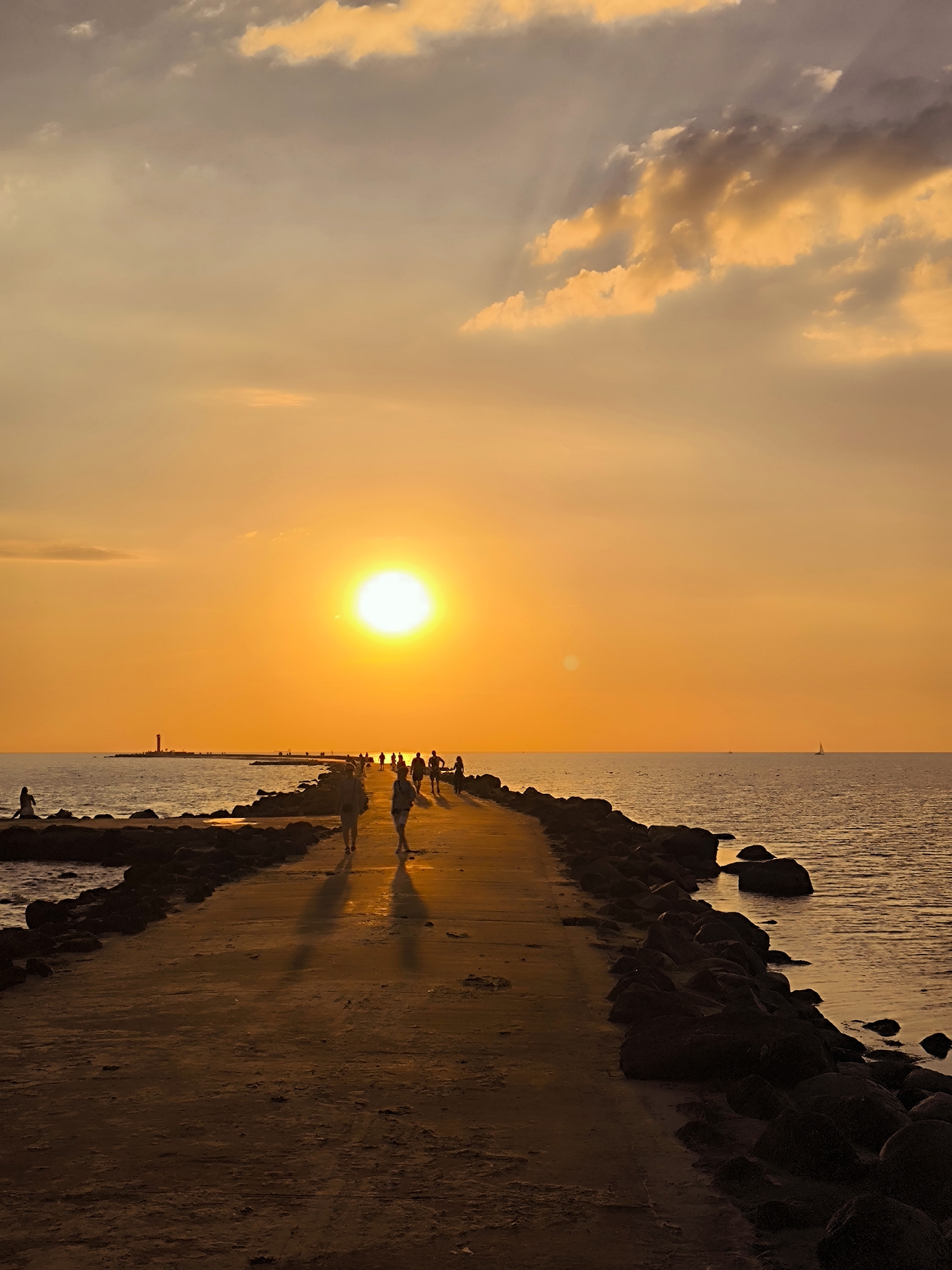 This image depicts a sunset over the Baltic Sea, viewed from a pier or breakwater. In the distance, a red lighthouse stands on the shore. Several people are walking along the pier, silhouetted against the vibrant orange sky. The water is calm, with a sailboat visible on the horizon. The pier is made of concrete and lined with rocks on the seaward side. The scene appears to be the Daugava River estuary, leading into the Baltic Sea, a common area for recreation in Riga, Latvia. The lighting suggests late afternoon or early evening.