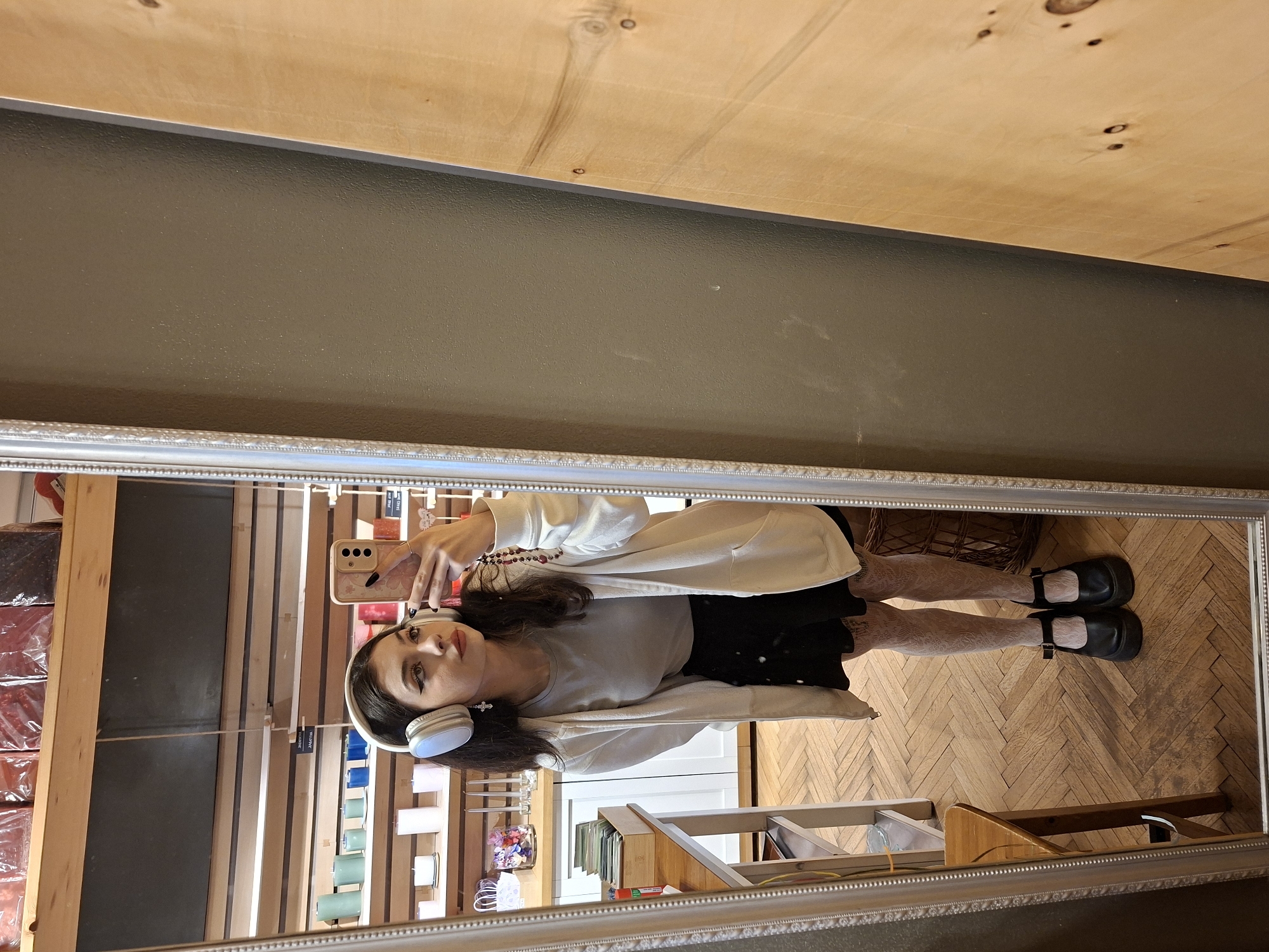 A photograph taken indoors shows a mirror reflection of a woman in an interior space. The woman is wearing a gray shirt, a black skirt, a white cardigan, patterned tights, and black Mary Jane shoes. She has white headphones around her neck and is holding a beige cellphone in her left hand, taking a photo of herself. The background includes a display of candles and other merchandise, suggesting a retail environment. A wooden floor with a herringbone pattern is also visible. The reflection suggests the photo was taken in a shop. Location information is not discernible.