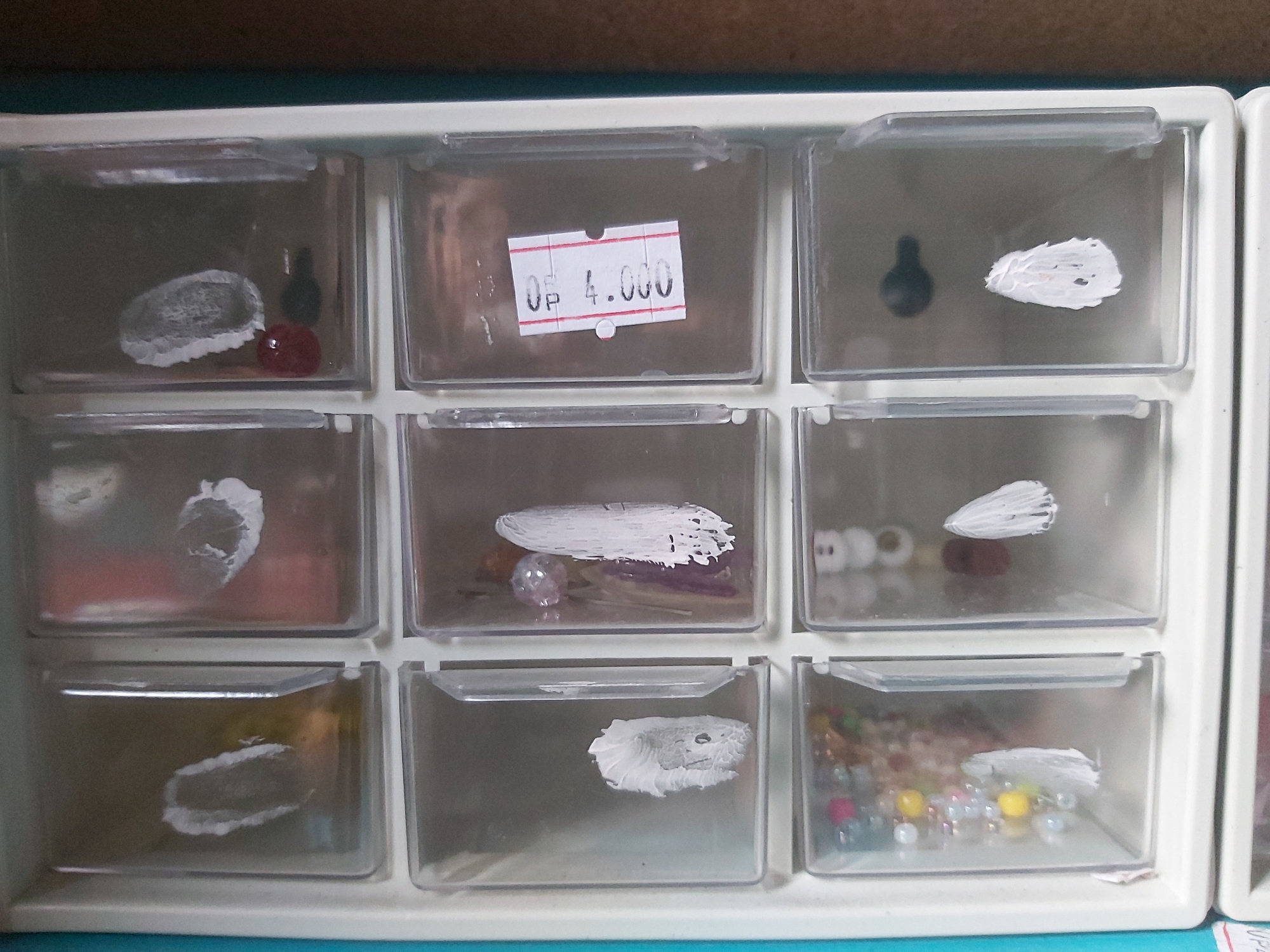 The image shows a white plastic organizer with multiple transparent drawers, each containing various small items. The organizer appears to be placed on a shelf or table with a wooden surface visible above it.
The top row of drawers contains:
* **Left drawer:** A grey, oval-shaped object with white markings, possibly some sort of stone or bead, with a small red spherical object and a black object, possibly a bead, to its right.
* **Middle drawer:** A white price tag with red lines and black text that reads "0 P 4.000".
* **Right drawer:** A black conical object on the left and a white smudge, possibly paint, on the right.
The middle row of drawers contains:
* **Left drawer:** A white smudge on a greyish object.
* **Middle drawer:** A clear spherical object with some colored items and a white smudge on top.
* **Right drawer:** Several small white and red spherical objects, along with a white smudge on the right.
The bottom row of drawers contains:
* **Left drawer:** A greyish object with white markings.
* **Middle drawer:** A white smudge on a grey surface.
* **Right drawer:** A collection of colorful beads, including clear, yellow, pink, and red ones, with a white smudge on the right.
The overall scene is one of organized storage of craft supplies or small collectibles. There are no people visible in the image. The environment is indoors, with artificial lighting. The location is described as Ploso, Indonesia.