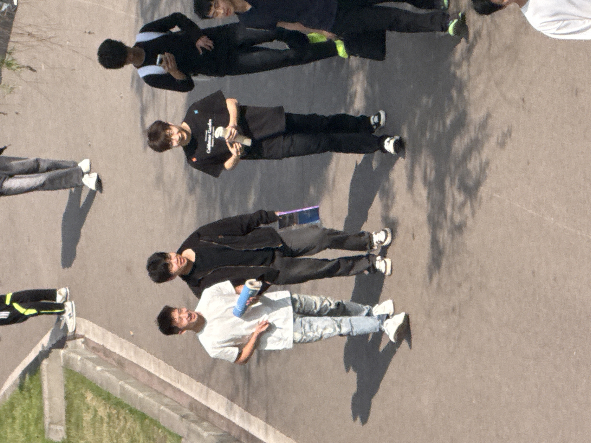 In Jiangjin, China, a group of people are walking on a paved surface. The individuals are dressed in various clothing styles, including sportswear, casual outfits, and a white t-shirt and patterned jeans. They are holding items such as a phone, a container, and a book. The environment consists of concrete, grass, and the shadows of trees.