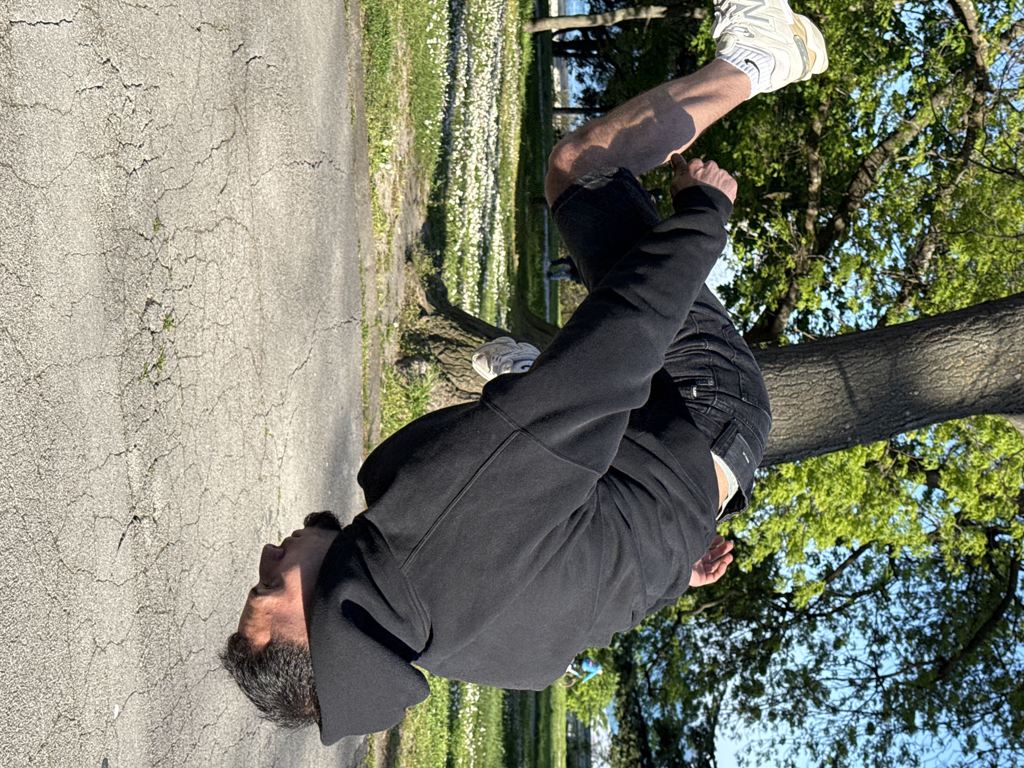 A man is photographed outdoors in Varna, Bulgaria, performing a backflip. He wears a black hooded sweatshirt, dark denim shorts, and white sneakers. He is inverted, with his feet up and his head pointing toward a cracked asphalt surface. The background includes a grassy area with white flowers, a tree trunk, and green leafy branches against a blue sky. The sun appears to be shining.