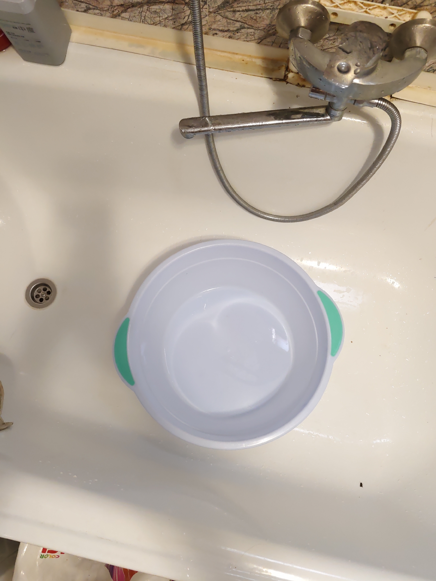 An overhead view of a white bathtub shows a white basin with green handles positioned slightly right of center. To the left of the basin is a silver drain. In the background, a chrome faucet with a flexible hose is mounted on the tub's edge. The background wall is tiled with a marble-like pattern. The environment appears to be a bathroom. The image is located in Rostov-on-Don, Russia.