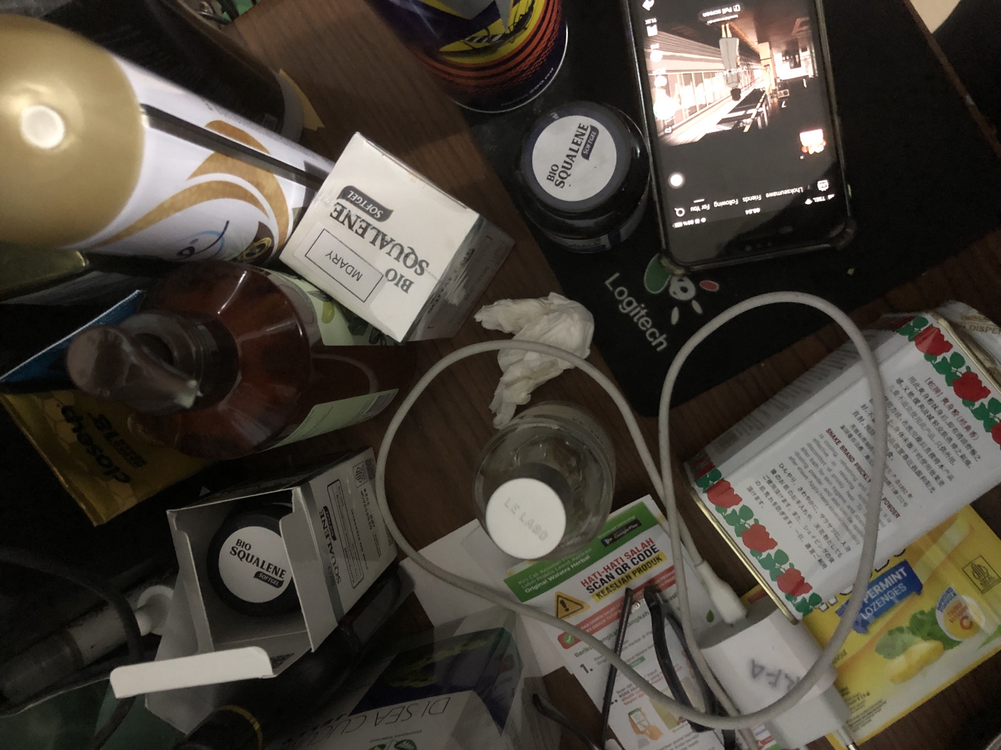 The image depicts a cluttered tabletop surface with various personal care products and electronic devices. In the center, a white box with the label "BIO SQUALENE SOFTGEL" is partially open, revealing a black circular container inside with the same branding. Adjacent to it is a larger brown bottle with a pump dispenser, and a tall, gold-capped bottle with a white label featuring a golden droplet design. A small, clear glass bottle with a white cap is also visible, along with crumpled white tissue paper. A grey charging cable snakes across the surface, connected to a white power adapter. To the left, a portion of a white box labeled "DI SEA CU" is present. Further to the right, a cylindrical object with colorful stripes, resembling a can, is partially visible. At the bottom of the frame, a smartphone displaying a video interface is resting on a black mat with a "Logitech" logo. In the background, dark, out-of-focus objects suggest a chaotic environment. The scene does not provide any specific visual cues or context to determine its location in Lhokseumawe, Indonesia.