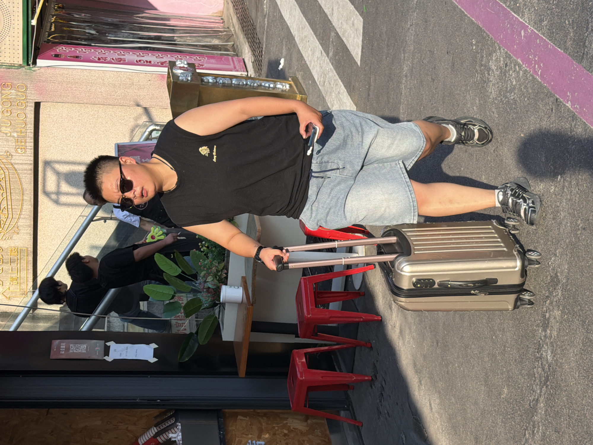 The image depicts a person with short hair, wearing sunglasses, a black sleeveless shirt, and denim shorts, pulling a gold suitcase with their right hand. The person is walking on a paved street with white diagonal stripes, indicative of a pedestrian crossing. The scene appears to be outdoors, likely in a city environment. In the background, there are buildings with signage, a staircase, and some red stools. A person in a black shirt is visible on the stairs. The lighting suggests it is daytime, with strong shadows cast on the ground. The environment appears to be Yuzhong District, China, as indicated by the signage and architectural style.