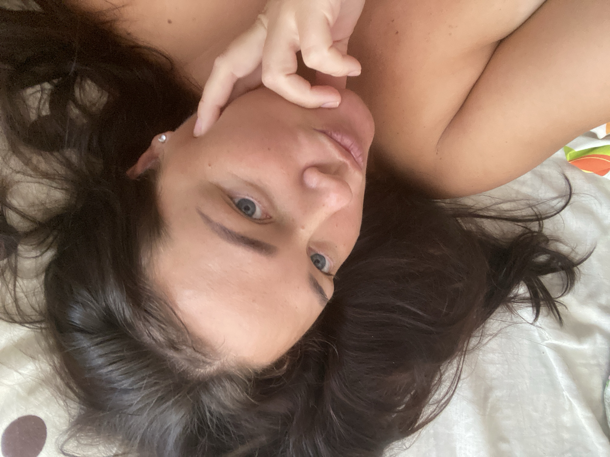 An overhead close-up shot shows a woman with dark hair and blue eyes lying on a white, textured surface. She is looking up towards the camera with her right hand touching her cheek and mouth area. A small diamond stud earring is visible in her right ear. The background shows a light-colored, textured surface, possibly a bedsheet, with subtle circular patterns. The lighting suggests an indoor environment. There is no identifiable information within the image to indicate the specific location in Sochi, Russia.