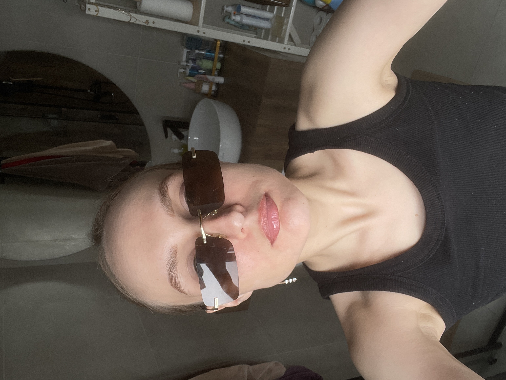 A woman wearing sunglasses and a black tank top is centered in the frame, angled towards the right, suggesting a selfie. The background features a bathroom setting with a white sink and mirror visible. Shelves above the sink hold various toiletries and household items. The floor and surrounding surfaces appear to be tiled. There is no information to confirm the location in the city of Podshivalovo, Russia.