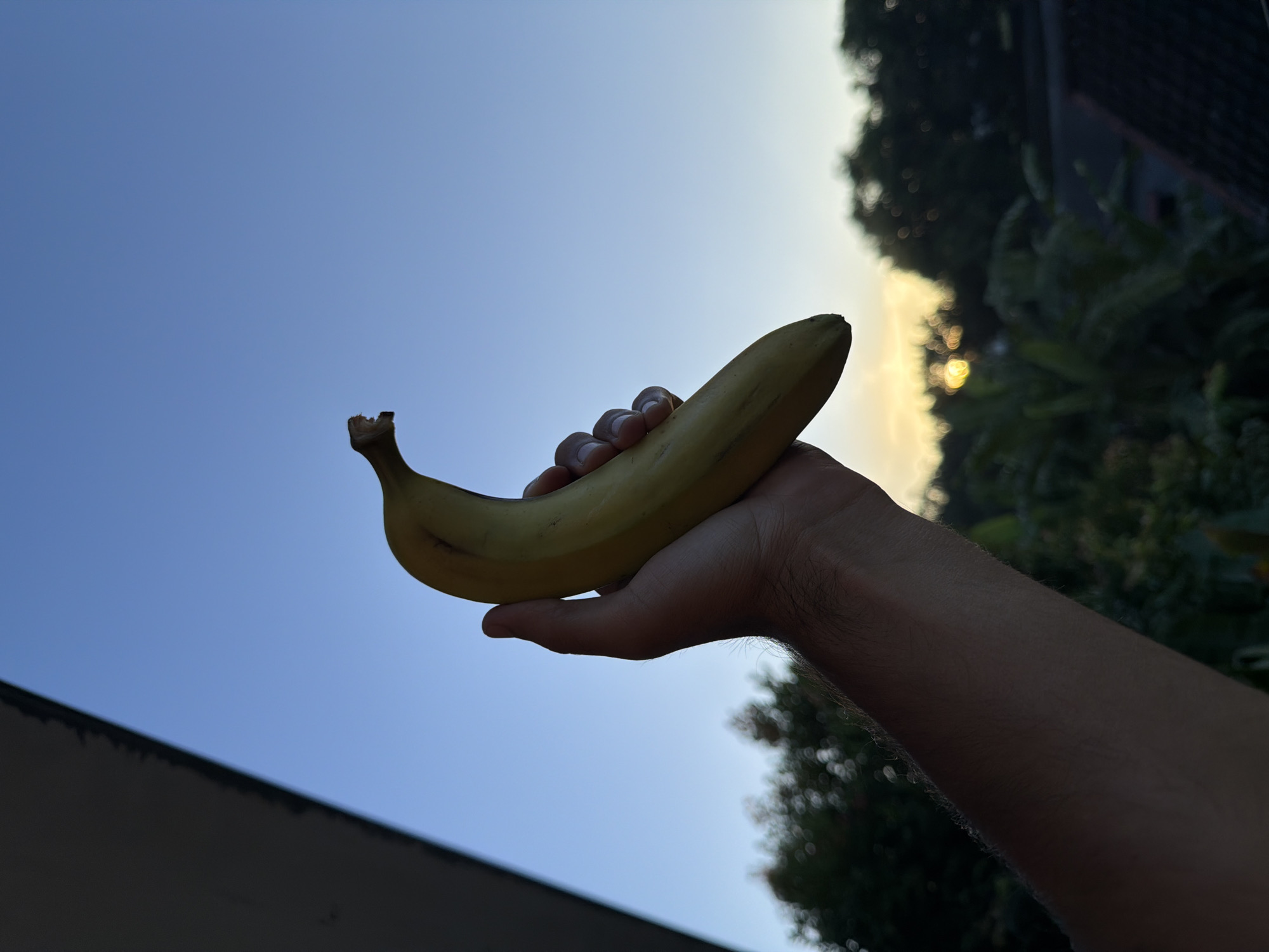 A person's arm and hand are extended, holding a yellow banana against a backdrop of a blue sky. The background also includes some trees and foliage. The photo was likely taken in a location with tropical vegetation, which aligns with North Kuta, Indonesia, but precise geolocation is impossible without additional information.
