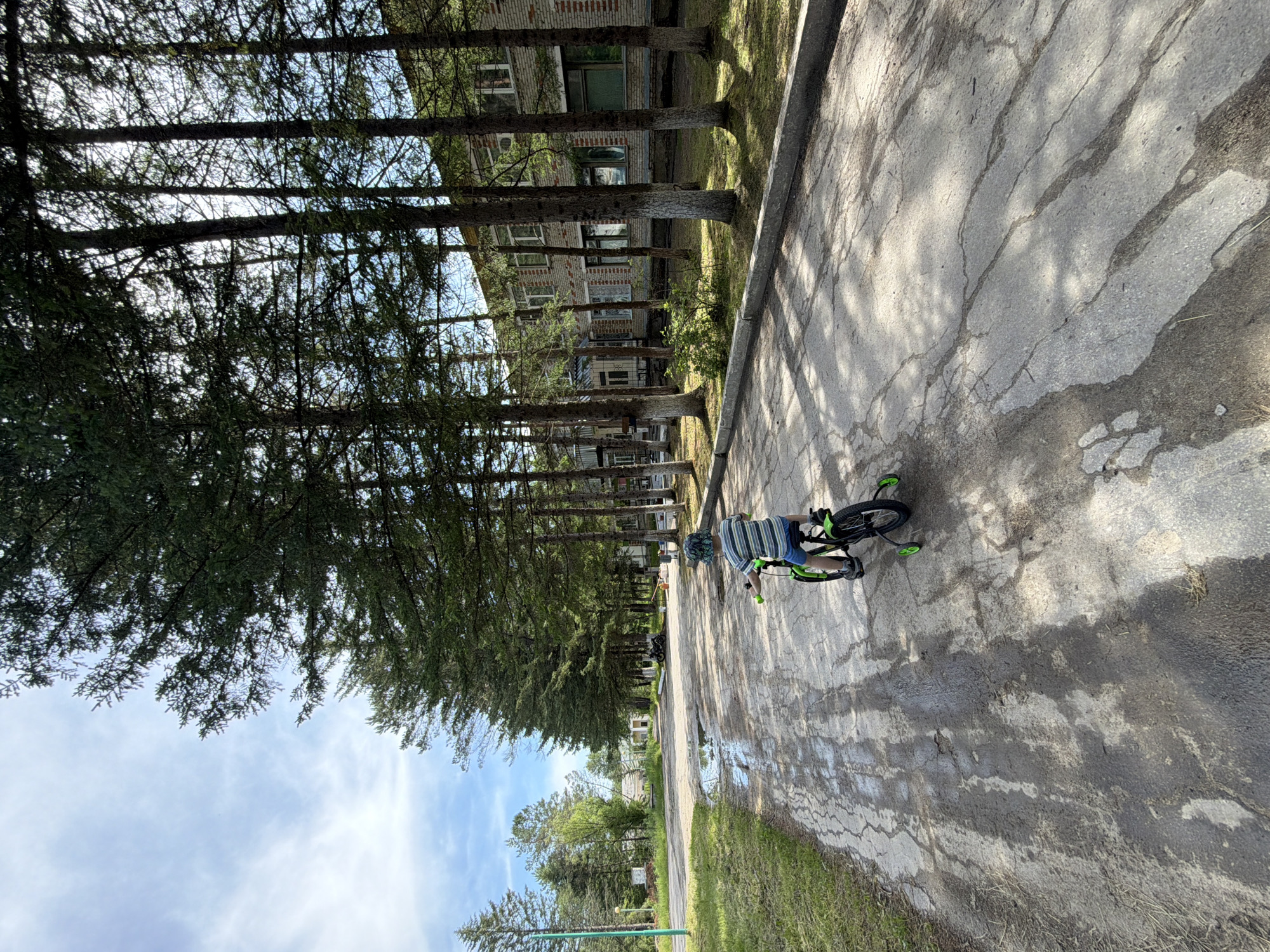 In Kul'dur, Russia, a child is riding a green bicycle with training wheels on a paved road. The child, facing away from the camera, wears a striped shirt and shorts, and a hat. The road is cracked and wet in patches, reflecting the sunlight and shadows of the tall evergreen trees lining the right side of the road. In the background, a multi-story brick building with numerous windows is visible through the trees. The sky is partly cloudy with patches of blue. To the left of the road, there is a grassy area with a tall green pole. Further down the road, other buildings and trees are visible. The overall scene depicts a quiet, somewhat weathered, outdoor environment.