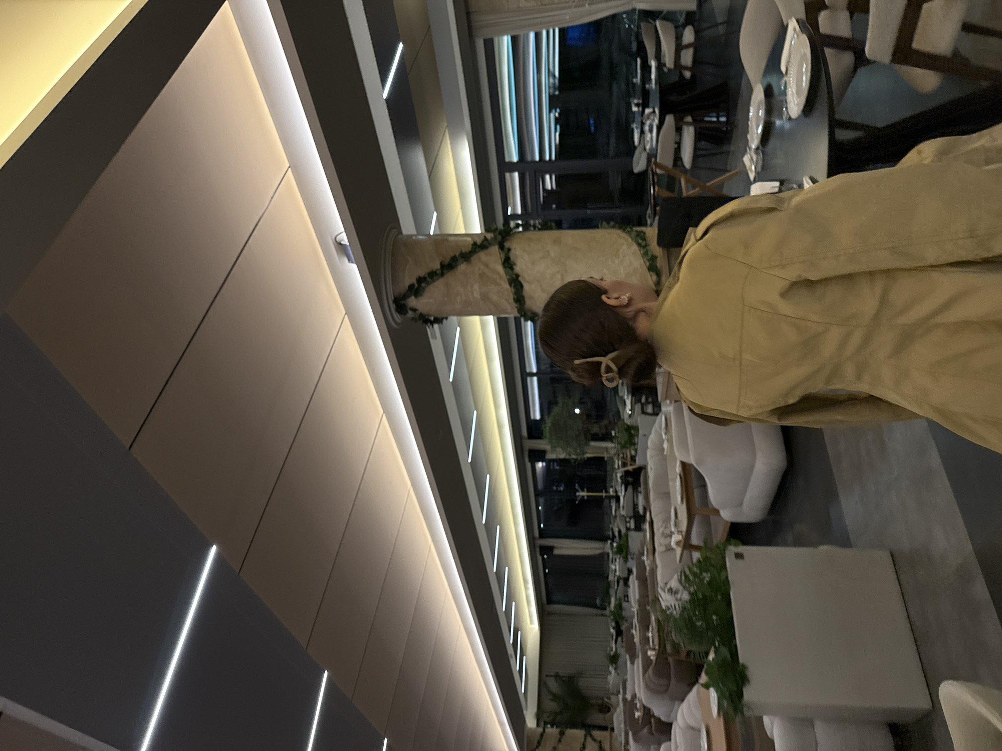 Here is a description of the image:
The photo depicts the interior of a restaurant or dining establishment in Tsakhkadzor, Armenia. A person wearing a tan jacket stands near a decorative column adorned with greenery. White upholstered chairs and tables are arranged in a dining area, and there is an abundance of white seating throughout the space. Large windows and architectural lighting are visible in the background.