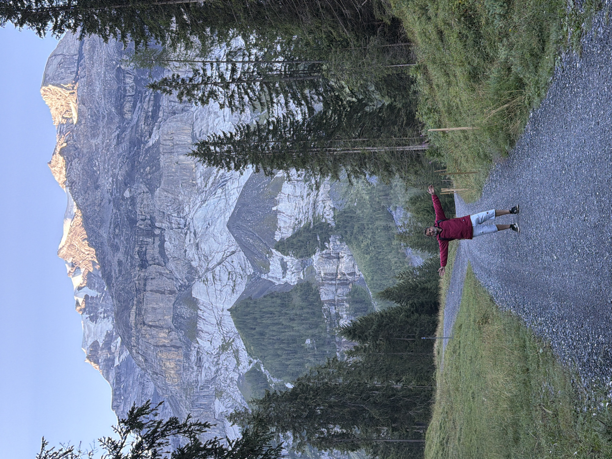 A man stands on a gravel path in Kandersteg, Switzerland, with his arms outstretched. He wears a red jacket and light-colored shorts. Behind him, a steep mountain face, partially covered in snow and ice, rises dramatically against a clear blue sky. Evergreen trees line the sides of the path and the lower slopes of the mountain. The sun illuminates the upper reaches of the mountain, casting a warm glow on the rocky terrain. The path curves to the left, leading into a forested area.