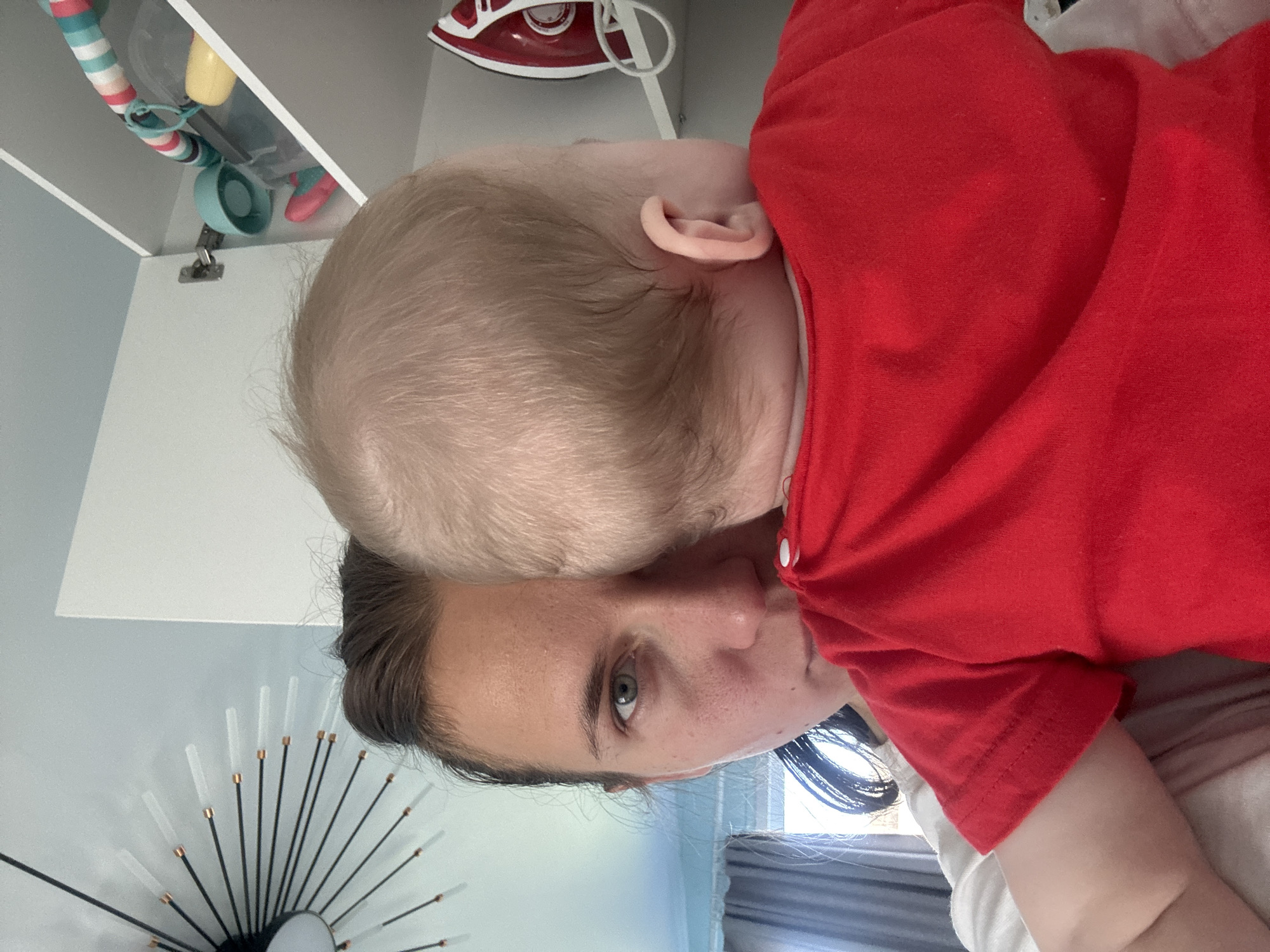 A woman with dark hair and fair skin is holding a baby wearing a red shirt. The baby's head is close to the woman's face. In the background, there is a white cabinet with various items, including an iron and some toys. A decorative sunburst mirror is also visible on the wall. The scene appears to be indoors. There is no information available to determine the location is in the city of Раздолье, Russia.