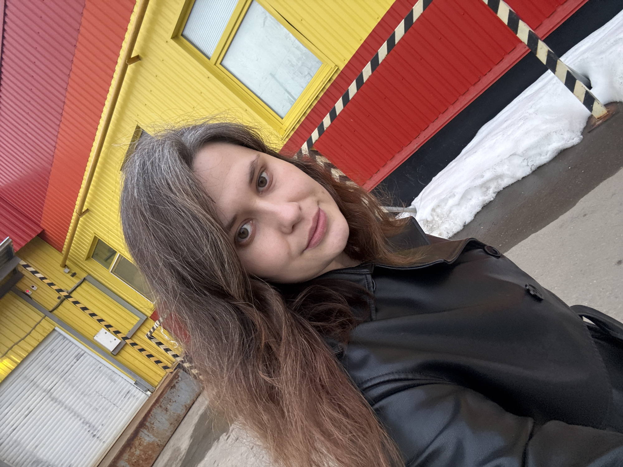 A female subject, estimated to be in her late 20s to early 30s, is depicted in a diagonally angled, medium-close shot against the exterior of a building in Chaikovsky, Russia. She has brown eyes, a slight smile, and long, multi-toned brown hair with visible grey streaks. She wears a black faux leather jacket with multiple buttons visible. The background is a structure composed of corrugated metal panels, predominantly painted in sections of vibrant yellow and red. A rectangular window with a yellow frame is visible in the upper-right portion of a yellow wall section. Below this, a red wall section features a black and yellow striped hazard pattern. Adjacent to the building's base on the right, accumulated white snow rests on wet, dark pavement. To the lower left, a large white corrugated roll-up door is set into a yellow wall, which also displays black and yellow hazard stripes and a vertical yellow pipe. Rusting is present on a lower metallic structure to the right of the roll-up door.