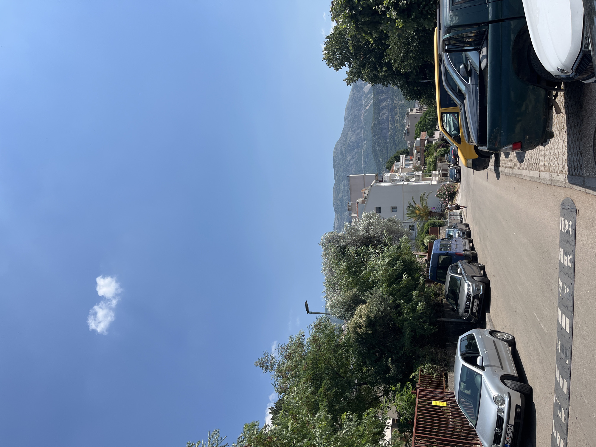 A street scene in Budva, Montenegro, under a clear blue sky with two small white clouds. The street is lined with parked cars, including a silver Volkswagen Polo, a gray SUV, a blue van, a yellow van, a dark-colored pickup truck, and other vehicles. Buildings with white facades and balconies are visible in the background, along with lush green trees and a distant mountain range. A speed bump is present on the road in the foreground.