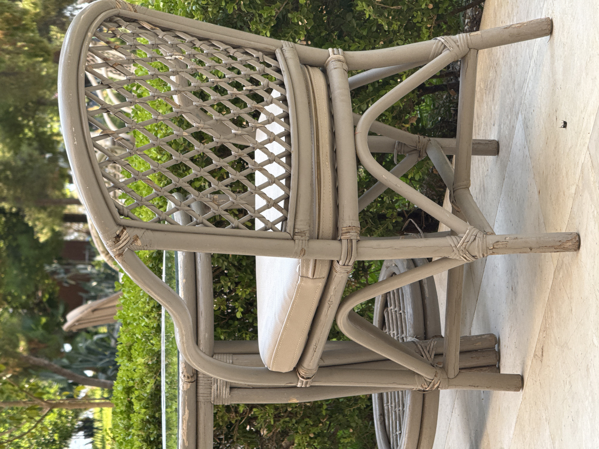 A light grey, weathered rattan chair with a light-colored seat cushion sits on a light-colored tiled patio. The chair is intricately woven with a diamond pattern on the back. Behind the chair is a dense hedge of green bushes, and further back, blurred trees and foliage are visible. The setting appears to be outdoors, possibly a garden or courtyard area in a warm climate. There are no people present in the image.