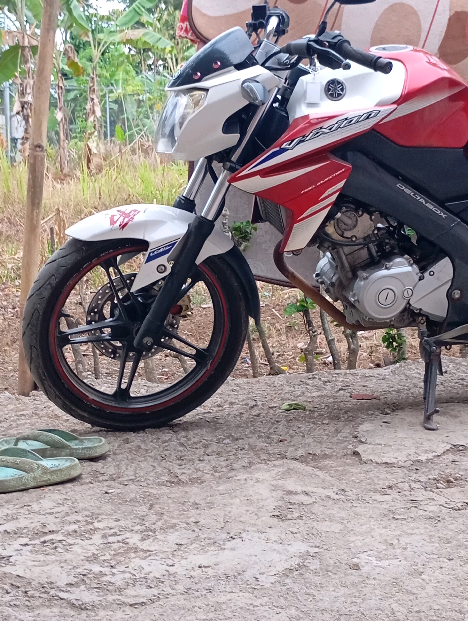 The image shows a red and white Yamaha motorcycle parked on a dirt road in a rural area, possibly Muncang, Indonesia, as indicated by the surrounding vegetation including banana trees and other foliage, and a wooden pole. The motorcycle is facing left, with its front wheel and part of the engine visible. A pair of light blue flip-flops are lying on the ground in front of the motorcycle's front wheel. The motorcycle has a decal on its front fender and the word "J-XION" along its side fairing, along with "DELTABOX" branding. The motorcycle is equipped with a kickstand. The lighting suggests it is daytime.