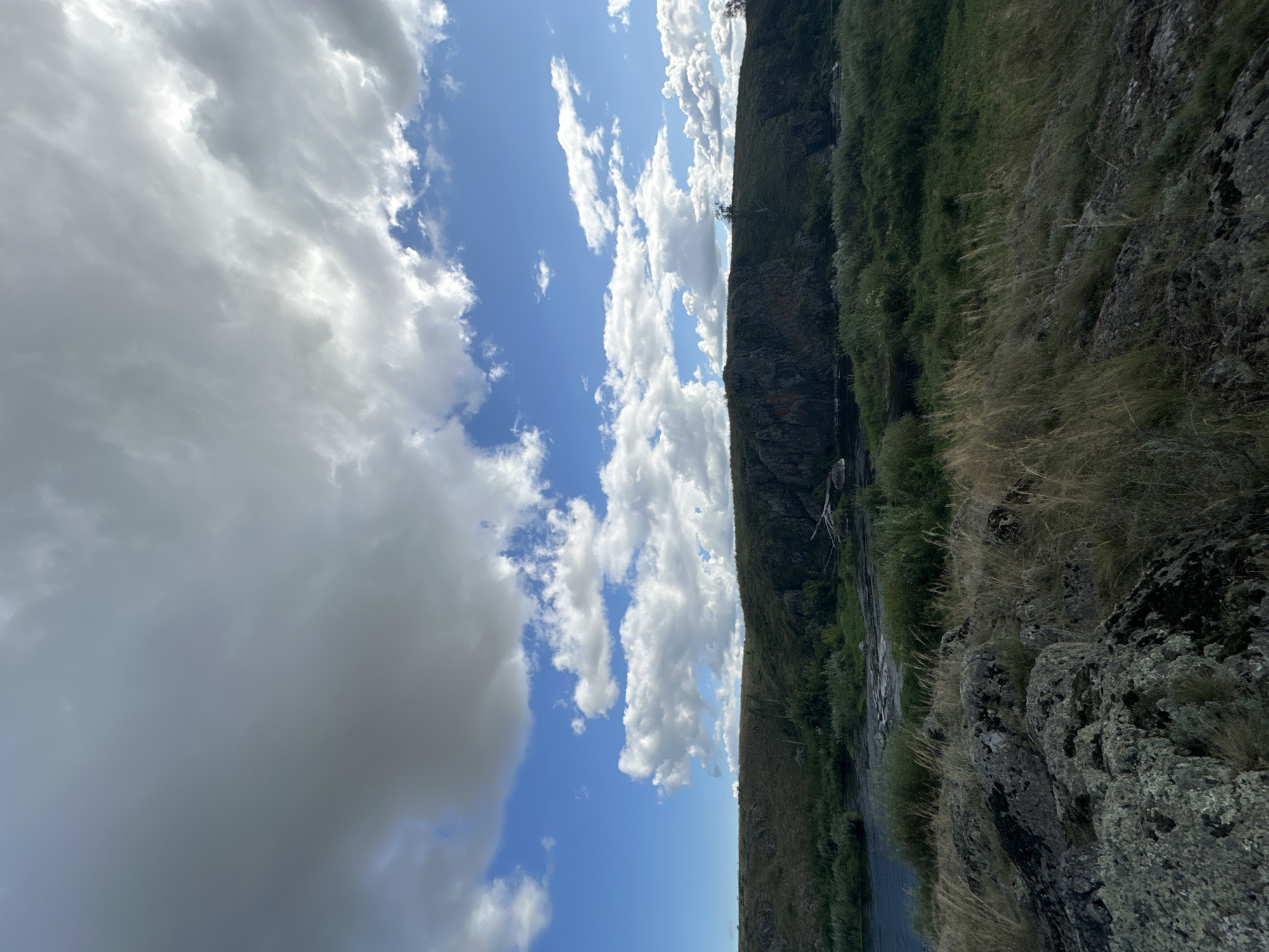 An outdoor, wide-angle shot captures a rugged, natural landscape under a dynamic sky. The foreground is dominated by large, grey rocks covered in patches of moss and lichen, with dry, golden grass growing between them. A river flows through the middle ground, its water appearing dark and calm, with a gentle ripple on the surface. On the far bank of the river, a steep, rocky hill rises, covered in dense green vegetation. The top of the hill is flat and appears to be a plateau. The sky above is a brilliant blue, filled with a dramatic display of white and grey clouds, with some of the clouds casting shadows on the landscape below. The overall impression is one of untamed wilderness.