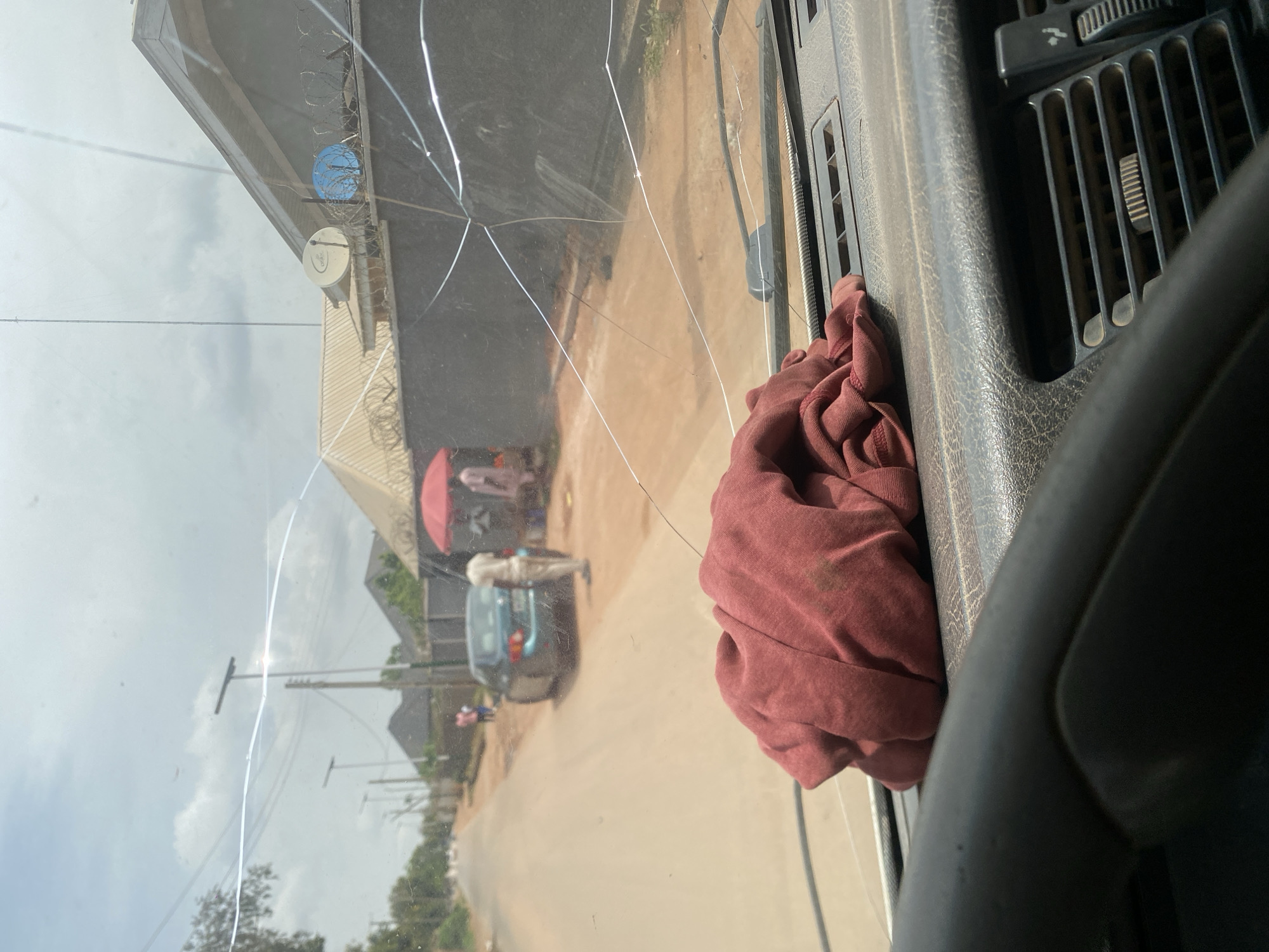 This image provides a view from inside a vehicle, looking through a cracked windshield towards a street scene in Rayfield, Nigeria. A crumpled red cloth rests on the dashboard in the foreground. The street is unpaved and dusty, with houses lining one side. Several people are visible on the street, including one standing near a blue car and others near a stall with a red umbrella. Streetlights are present along the road. The sky is overcast. Satellite dishes are visible on the roof of one of the houses.