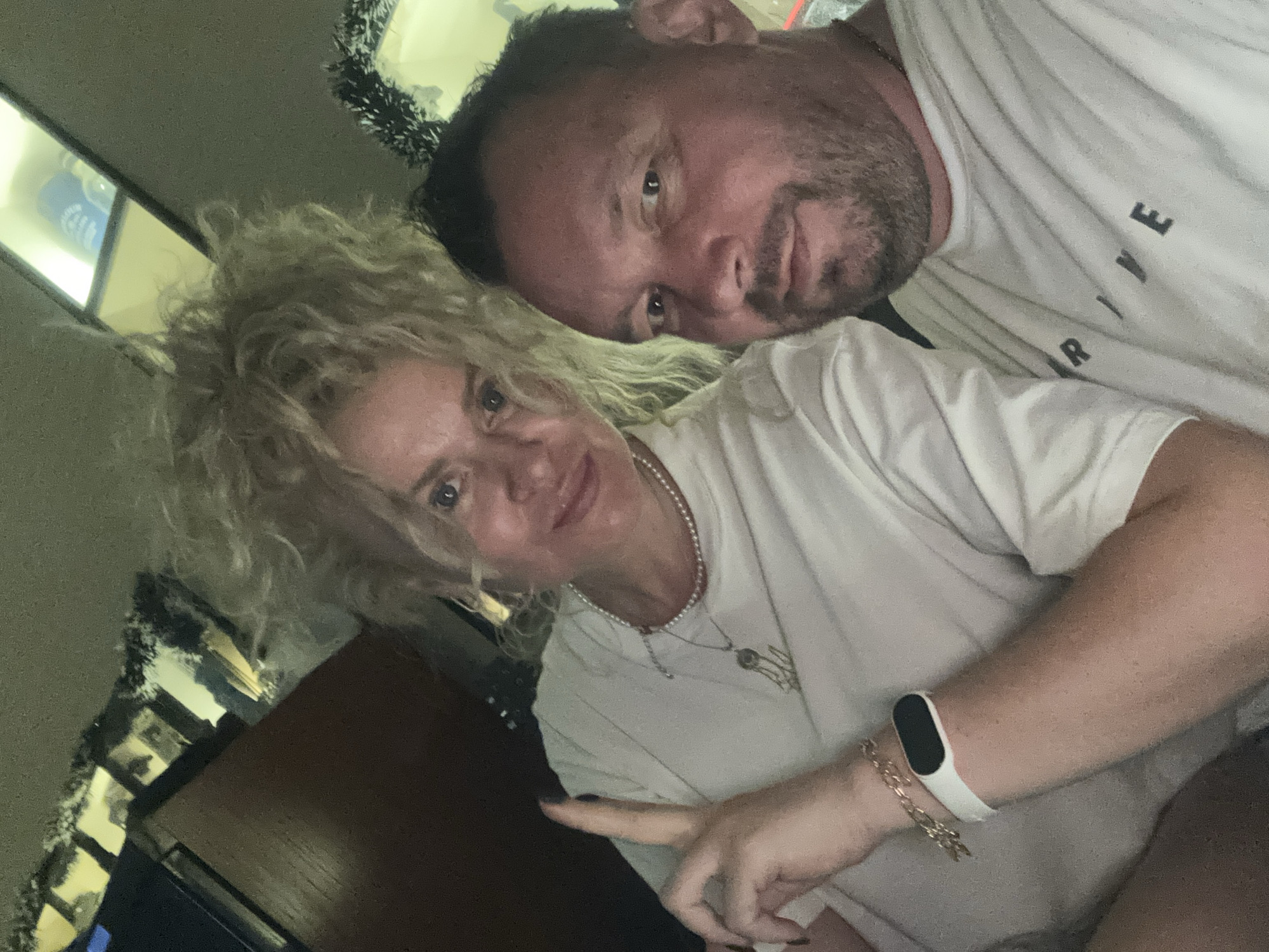 In a close-up, selfie-style shot, a man and a woman are positioned in the foreground. The woman, with voluminous blonde curly hair, is on the left, her face angled towards the camera with her left hand raised, index and middle fingers extended in a peace sign. She is wearing a white t-shirt, a pearl necklace, and a bracelet. The man, with short dark hair and stubble, is on the right, also facing the camera. He is wearing a white t-shirt with the word "DRIVE" visible in black letters across the chest. They appear to be indoors, with a dark wooden surface and shelves with illuminated objects in the background. A string of white and black garland is visible to the right of the woman's head. The lighting suggests an indoor environment, possibly at night. The image does not provide any information to place it in the city of Nanhai District, China.