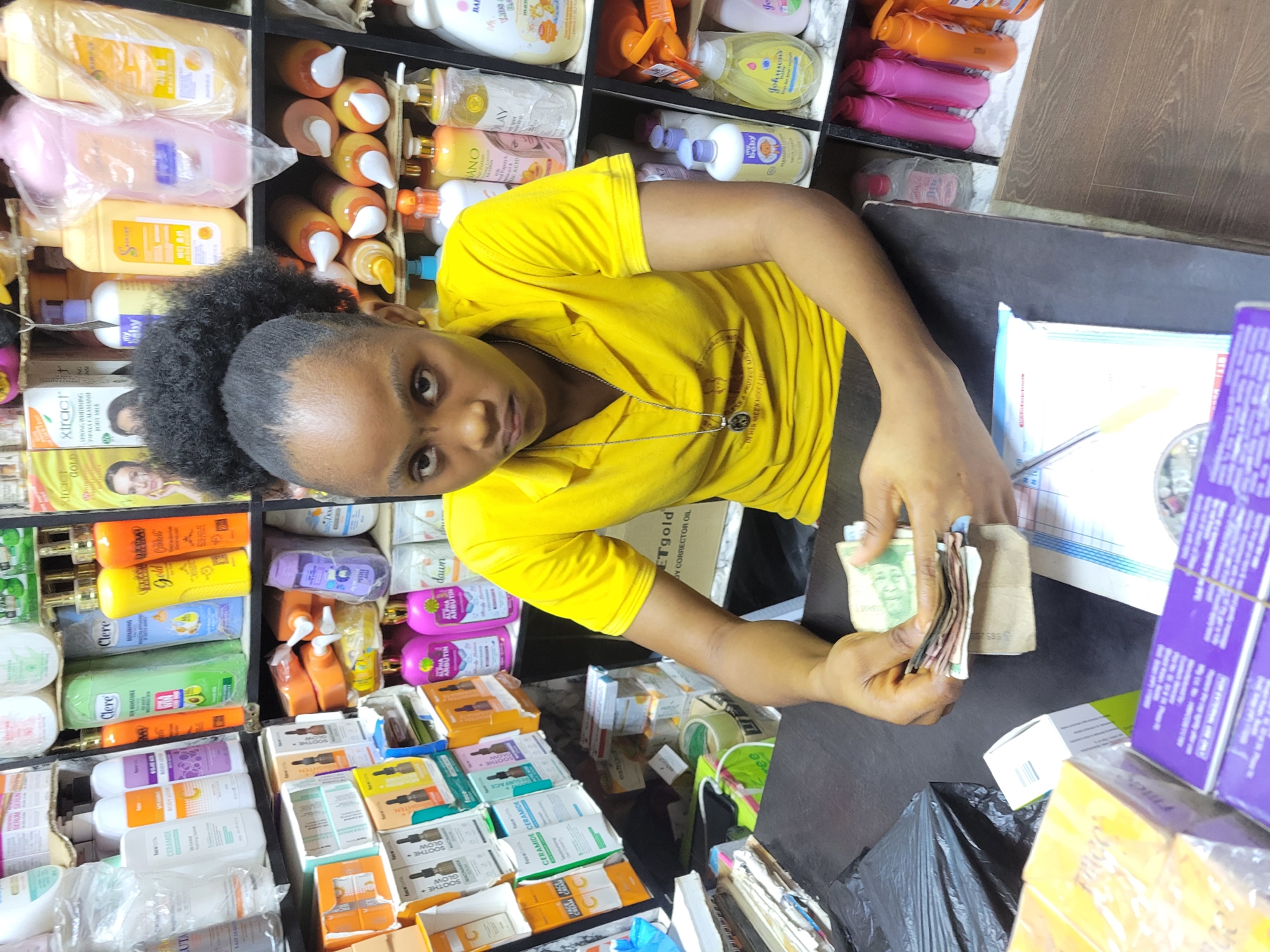 A woman in a yellow shirt stands behind a counter in what appears to be a small store in Onitsha, Nigeria. The store is filled with beauty products, lotions, and creams. The woman is holding a stack of Nigerian Naira notes in her hands and appears to be counting them. The counter is cluttered with paperwork, packaging, and a black plastic bag.