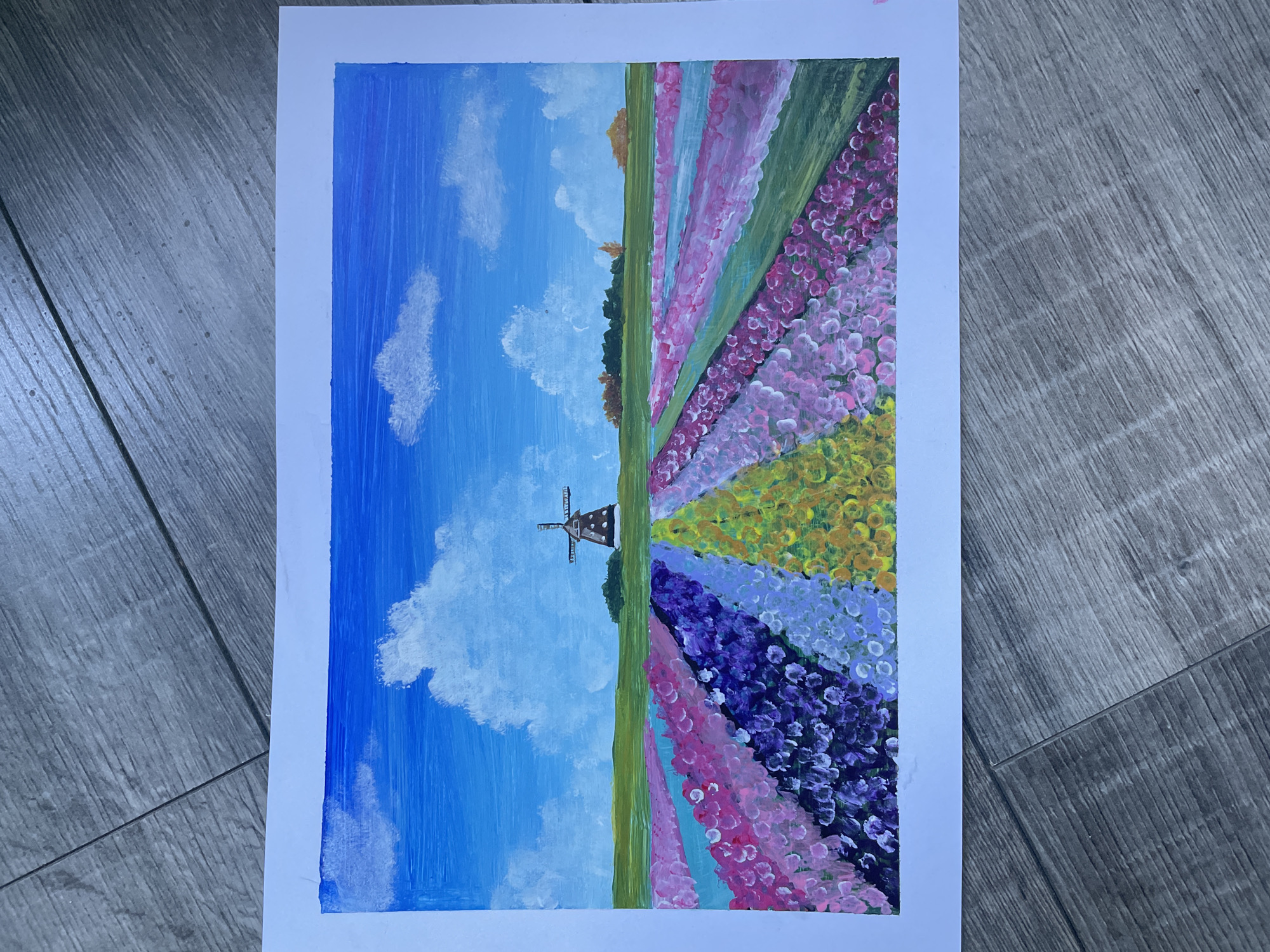 A painting depicts a colorful field of tulips under a bright blue sky with fluffy white clouds. In the distance, a traditional Dutch windmill stands against a backdrop of green hills and trees. The foreground is filled with rows of tulips in various vibrant colors, including pink, purple, yellow, and blue, creating a rainbow effect. The style of the painting is impressionistic, with visible brushstrokes that give texture to the flowers and sky. The painting is placed on a wooden surface, likely a floor, with visible wood grain and planks.