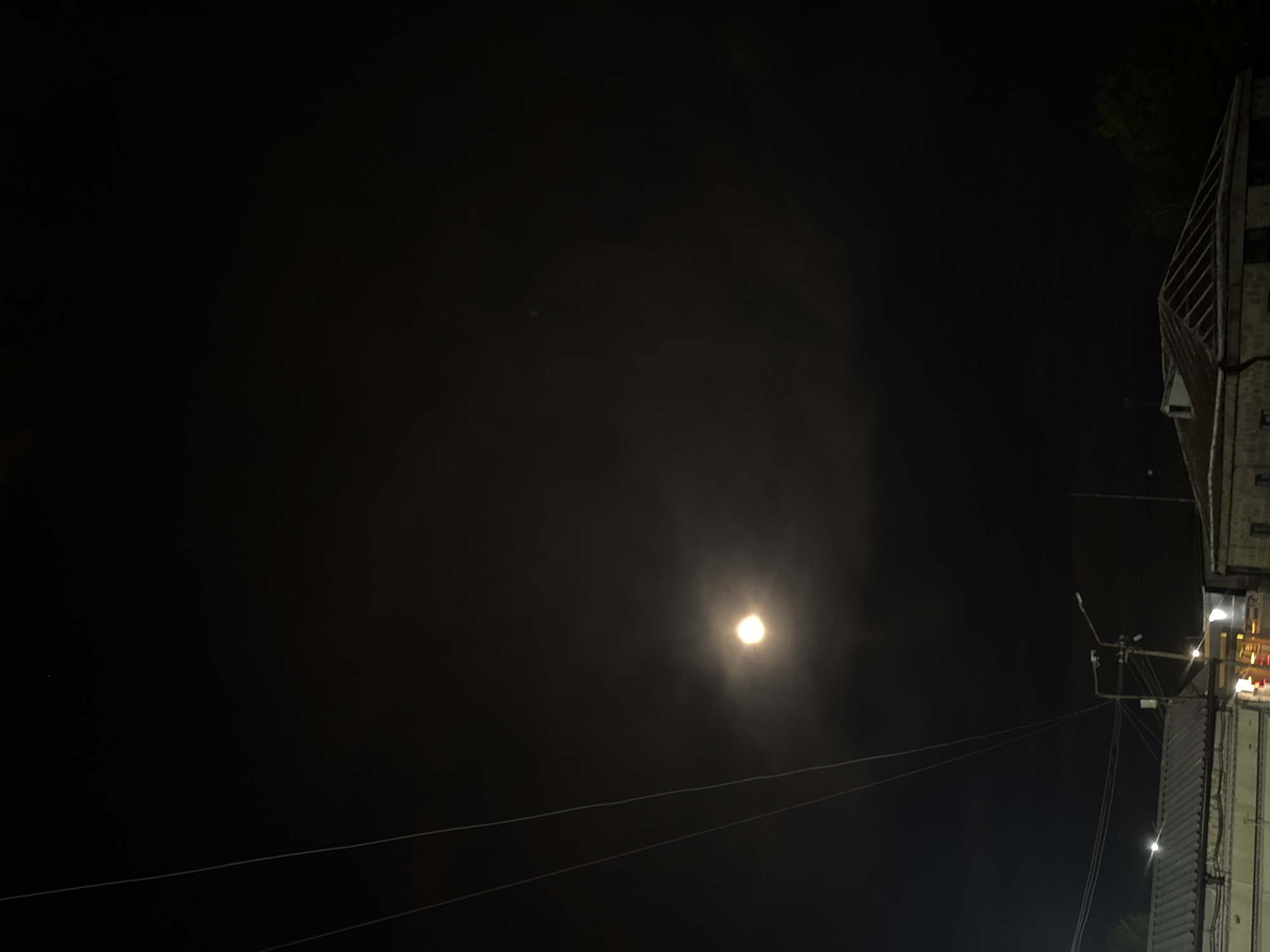 The image captures a nighttime scene dominated by a dark sky, likely in Yerevan, Armenia. A single, very bright light source, resembling the moon through a hazy atmosphere or lens flare, is prominently positioned slightly to the left of the center, emitting a soft, circular glow. Two thin, parallel lines, appearing to be power lines or utility cables, stretch across the lower portion of the frame, running horizontally from right to left.
On the far right side of the image, a tall building or structure is partially visible, extending vertically along the edge. This building exhibits a distinctive facade with pronounced vertical elements, possibly architectural louvers or textured panels. Several small, localized artificial lights are visible on or near the structure. A small section of dark tree foliage is discernible in the top right corner, above the building. The overall lighting suggests deep night, with the bright celestial body being the primary light source beyond the scattered artificial lights on the building. No people or discernible text are present in the frame.