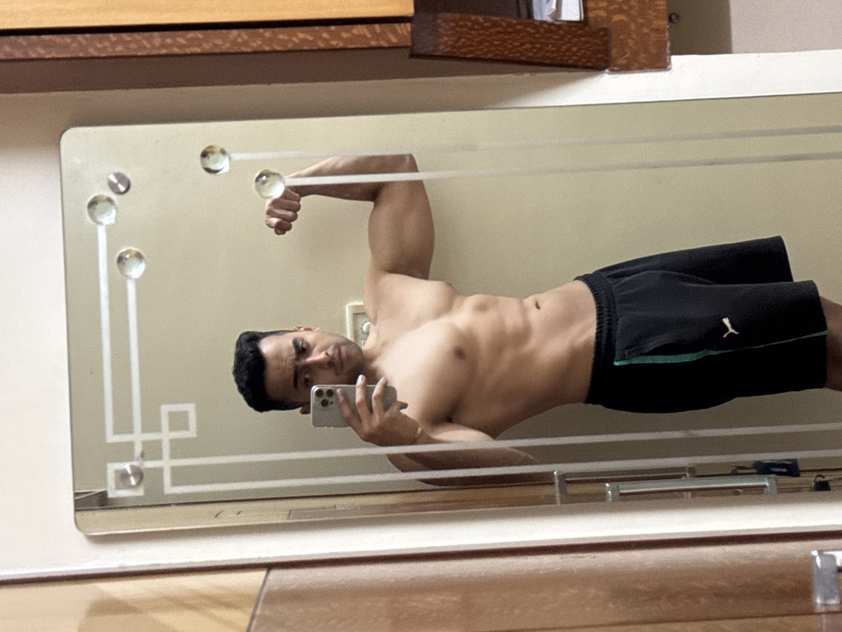 A shirtless man takes a selfie in a mirror. He is wearing black athletic shorts with a green stripe and is flexing his arm. The mirror has a vintage design with glass spheres and geometric patterns. The background includes a wooden floor and a cabinet. The man is holding a smartphone in his left hand, and his face is visible in the reflection.
