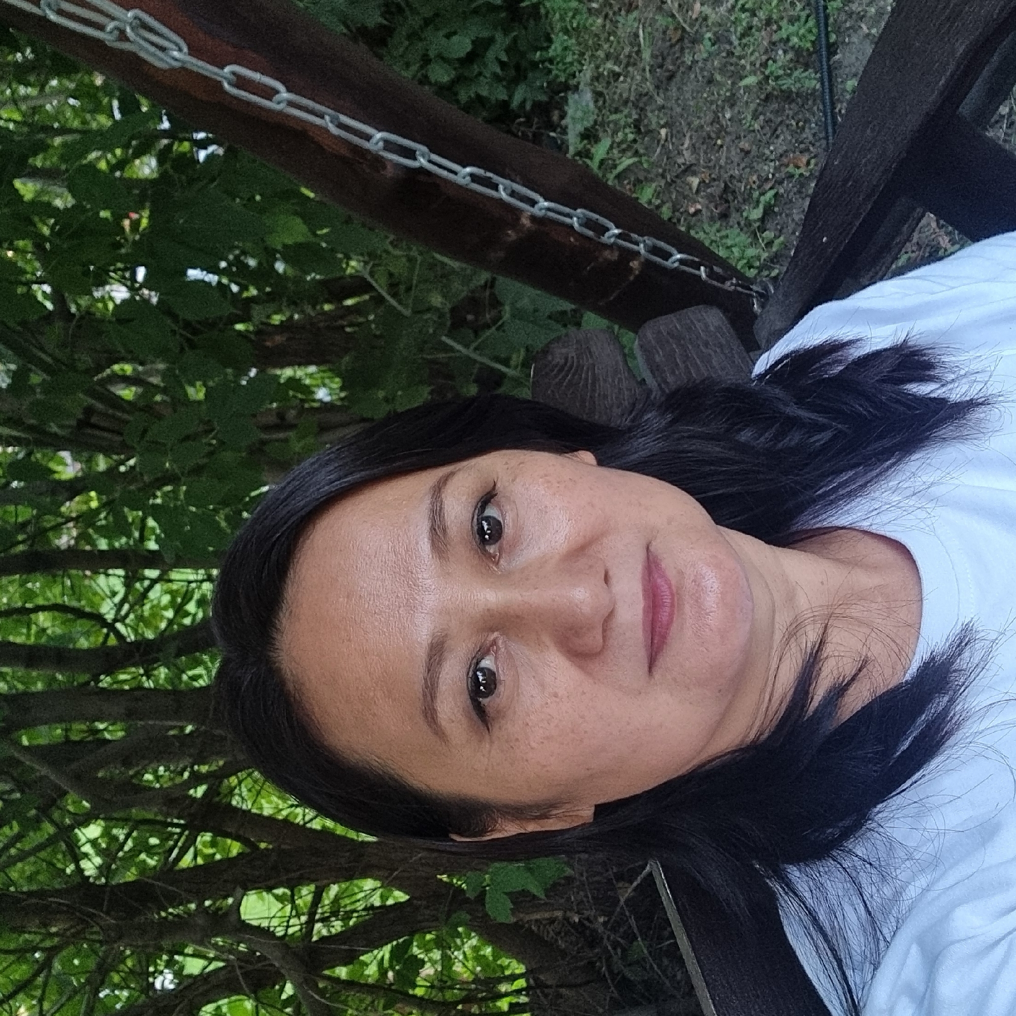 A portrait photograph features a middle-aged East Asian woman with dark hair, seated outdoors on a wooden swing. She is wearing a white t-shirt. The background consists of lush green trees and foliage. The woman's facial expression is neutral as she looks directly at the camera. The lighting suggests it is daytime. The scene is consistent with an outdoor recreational area or a garden. There are no specific landmarks or elements in the image that would allow for definitive identification of the location within the city of Aktobe, Kazakhstan.