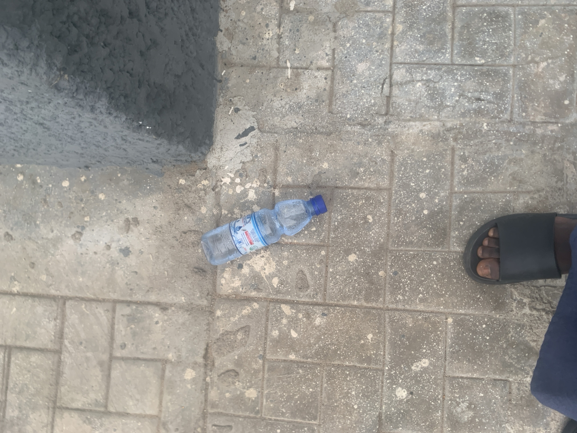 The image shows a plastic water bottle lying on a paved walkway. The walkway is made of rectangular concrete slabs with visible dirt and debris. To the right, a dark, rough-textured surface, possibly a painted wall or a large block, is visible. In the lower left corner, a person's foot wearing a black sandal is visible, along with a portion of their dark blue pants. The lighting suggests an outdoor, daytime setting. The location is identified as Jalingo, Nigeria.