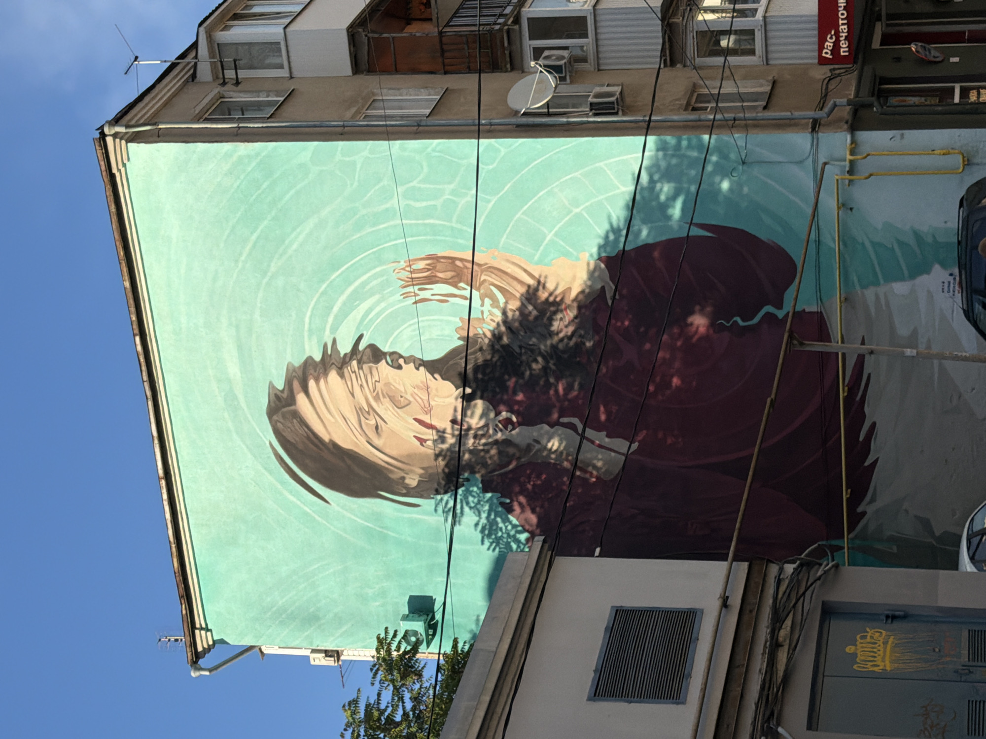 A low-angle, medium shot shows a mural on the side of a building in Rostov-on-Don, Russia. The mural depicts a person or figure whose image is distorted as though reflected in concentric ripples of water. The background of the mural is a teal or light turquoise color. The subject of the mural is mostly shades of brown, burgundy, and grey. Overhead, wires and pipes crisscross the scene, obscuring parts of the mural. The sky is bright blue. The adjacent building is visible.