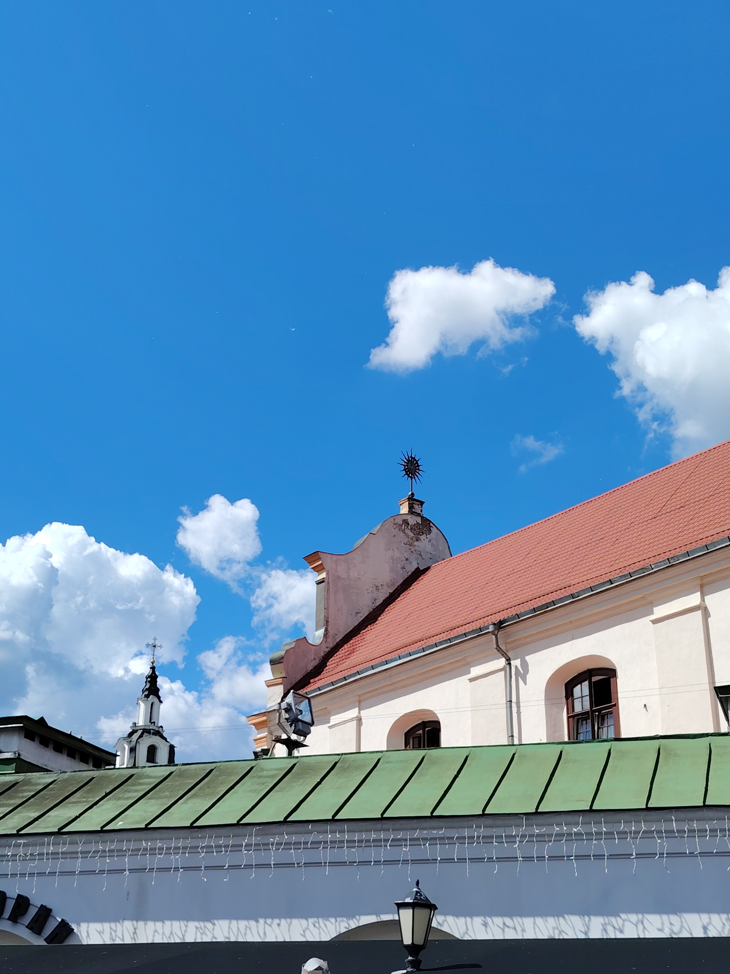 This outdoor daytime photograph captures an architectural street scene against a bright blue sky filled with scattered white cumulus clouds. The perspective is angled upwards, highlighting the upper sections of buildings and the expansive sky.
In the foreground, a white wall is topped by a distinctive green, slanted roof. White string lights, resembling icicle decorations, hang from the edge of this green roof. A black, ornate street lamp is partially visible at the bottom-center, and faint white text, including "OPAN", is discernible on a darker archway beneath the green roof.
Above the green roof, to the right, is a building with light-colored (beige or cream) walls and a prominent red-tiled roof. This structure features several arched windows with dark frames. A small architectural element on its upper section is adorned with a spiky, sun-like finial.
In the background on the left, a traditional church steeple rises, characterized by its white structure, a black dome, and a cross at its apex.
The scene is bathed in bright daylight, indicating clear and pleasant weather. A very faint crescent moon is subtly visible in the upper sky, along with some tiny, scattered specks, possibly dust or distant objects. The overall atmosphere is peaceful and serene, showcasing typical urban architecture under a vibrant sky, consistent with a location such as Minsk, Belarus.