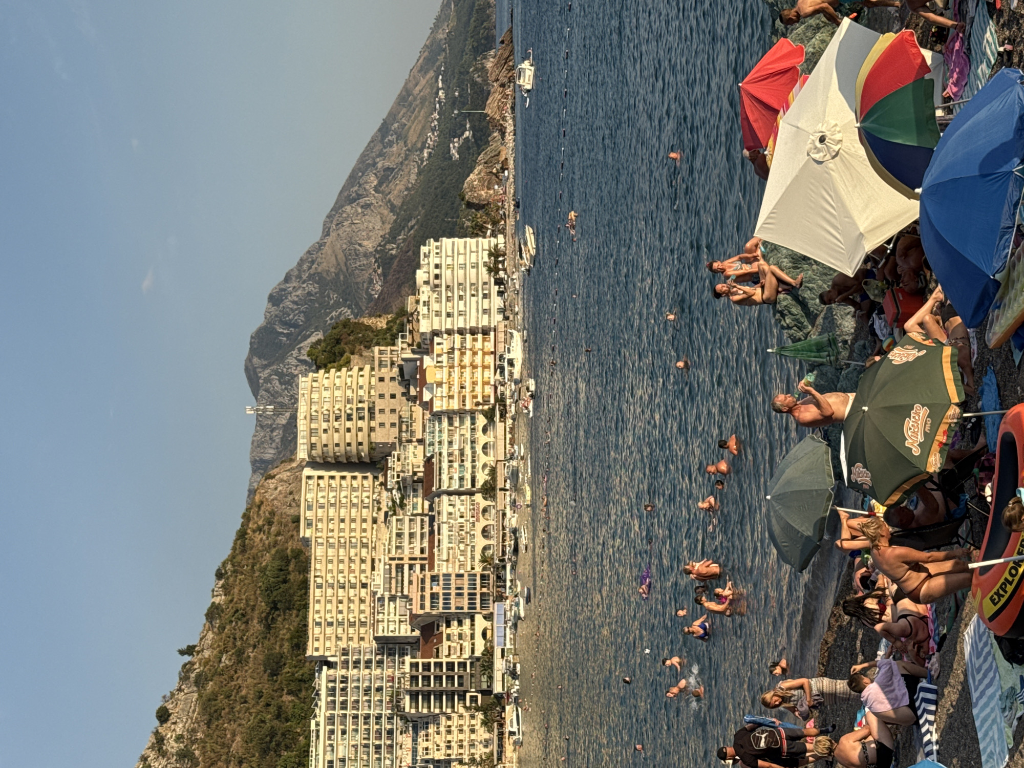 The image depicts a bustling beach scene in the city of Rafailovići, Montenegro, bathed in the warm glow of late afternoon sun. The foreground is a sandy beach populated by numerous sunbathers relaxing on towels and under umbrellas. In the water, a multitude of people are swimming and enjoying the sea. Behind the beach, a densely packed row of multi-story buildings, likely hotels and apartments, stretches along the coastline. The buildings are predominantly light-colored with numerous balconies, and they rise up the side of a steep, rocky mountain that forms the backdrop of the scene. Several boats are anchored in the calm, blue water between the beach and the buildings. The overall atmosphere is one of a vibrant summer holiday destination.