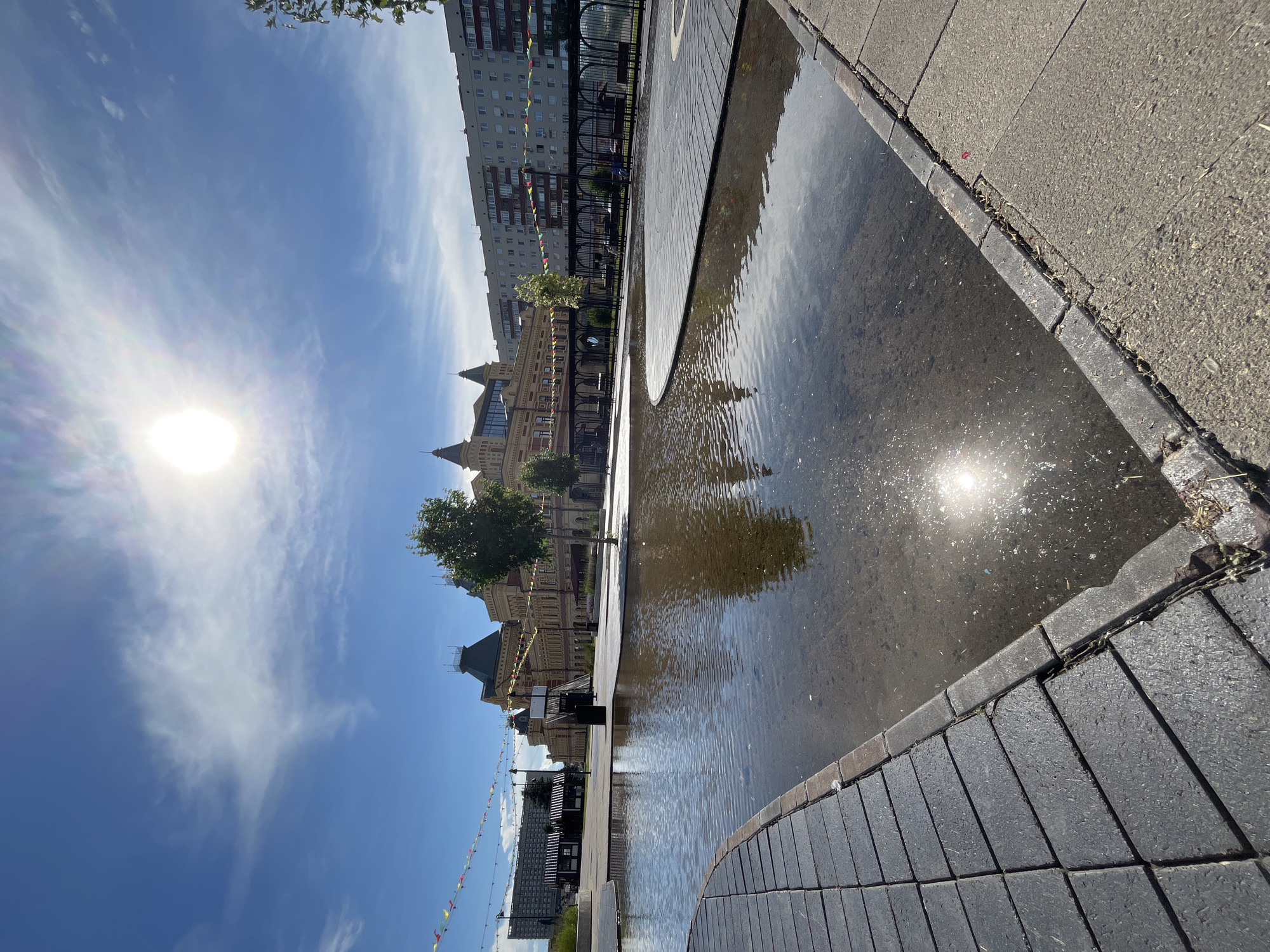 The image shows a sunny day in a city square in Nizhny Novgorod, Russia. In the foreground, a shallow, irregularly shaped body of water with ripples reflects the sun and the surrounding buildings. The water is bordered by a walkway paved with dark gray rectangular stones. In the middle ground, a tree stands on a paved area, and beyond that, a light brown, ornate building with a dark roof and multiple towers dominates the scene. Flags are strung across the area in front of this building. To the right of the ornate building, a modern multi-story apartment building is visible. The sky is blue with scattered white clouds, and the sun is bright and high in the sky. There are no people clearly visible in the scene. The environment appears to be an urban public space designed for leisure and recreation.
