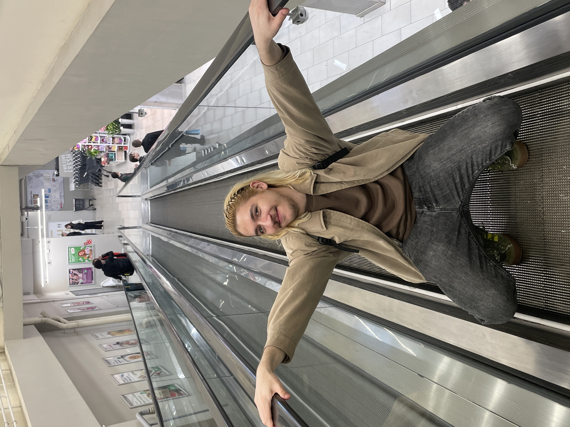 A young person with long blond hair and a brown headband is kneeling on a moving escalator, facing the camera, while holding onto both handrails. They are wearing a brown shirt, a beige jacket, and grey jeans with dark green shoes. The escalator is in a brightly lit indoor location, presumably a shopping mall or retail store. The background shows other people walking, product displays, and advertisements. Based on the store interior and the overall visual aesthetic, the location is likely in a large city, potentially Moscow, Russia.