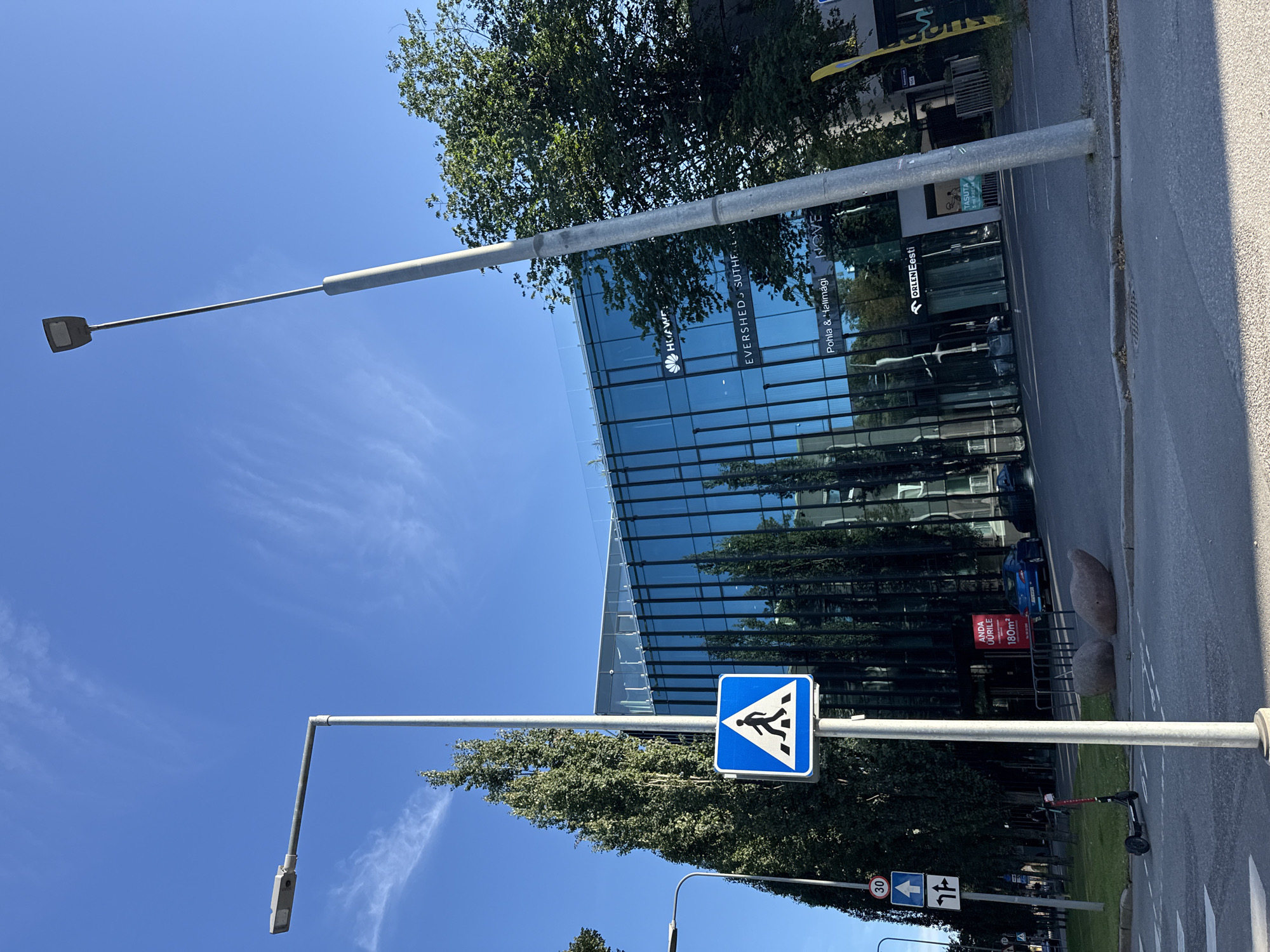 The image depicts a street scene in Tallinn, Estonia, dominated by a modern glass-fronted building. The building's facade reflects the bright blue sky and surrounding trees. Prominently displayed on the building are logos for "HUAWEI", "EVERSHED SUTHERLAND", "Pohla & Halimägi", and "NOVE". To the left, a blue pedestrian crossing sign is mounted on a pole, accompanied by traffic signs indicating speed limit (30), a straight-ahead arrow, and a left-turn arrow. Several tall, slender trees line the sidewalk in front of the building. A street lamp stands near the pedestrian crossing sign. In the foreground, a paved road with white pedestrian markings is visible, along with an electric scooter parked on the side. Two large, rounded stones rest on the grassy area beside the road. In the background, a blue car is parked in front of the building, and another car is partially visible behind it. The sky is clear blue with wispy white clouds.