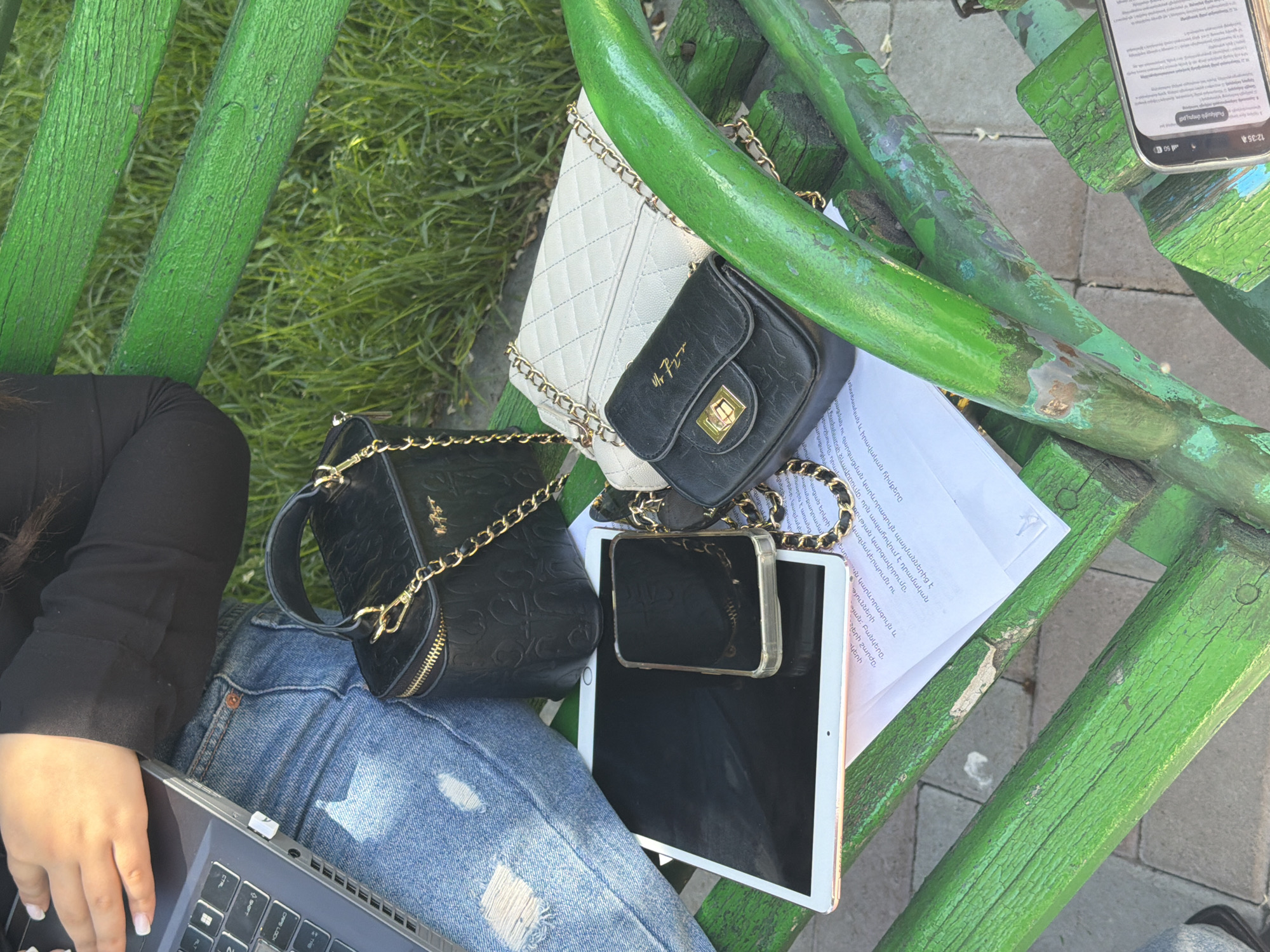 The scene is an overhead shot of objects and people on and around a green, wooden bench in Yerevan, Armenia. A person sits in the lower left corner, wearing a long-sleeved black top and blue jeans with rips. A laptop rests on their lap. Two purses, one larger black purse and a smaller white quilted purse with a black flap, sit on the bench. A smartphone in a transparent case is on top of the tablet. A white piece of paper with Armenian text is also on the bench. A phone is on the top right corner of the bench. The background includes patches of grass and paving stones.