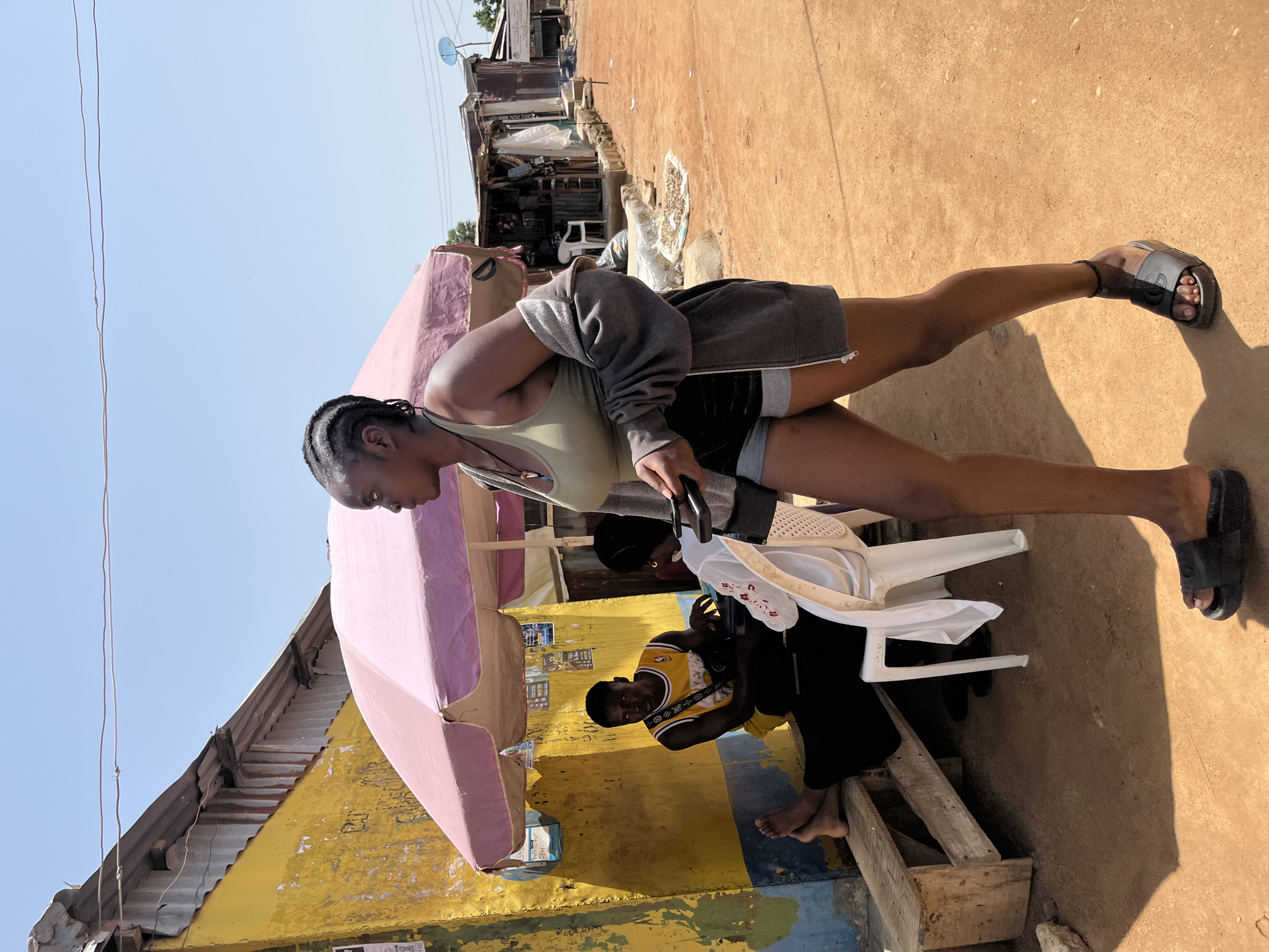 The image depicts an outdoor scene in Jos, Nigeria. The primary subjects are a woman and a man, with another person's legs visible in the background. The woman is standing, wearing a grey hooded jacket over a green tank top and black shorts. She has her hair in braids. She is holding a black object, possibly a phone or a remote. The man is seated on a wooden bench, wearing a yellow and black basketball-style jersey and black shorts. His legs are bare.
They are positioned near a stall with a large, pink umbrella providing shade. The stall is adorned with yellow and blue paint, and appears to be a small shop or market stall. Behind the stall, a row of similar structures forms a streetscape, with corrugated metal roofs and various goods visible within. In the background, a satellite dish is mounted on one of the buildings. The ground is covered in reddish-brown dirt, typical of the region.
The activity appears to be a casual gathering or perhaps related to business at the stall. The sunlight suggests it is daytime, and the clear sky indicates fair weather. Shadows are cast on the ground, further indicating a sunny day. The scene has a lived-in, informal feel, characteristic of a local market environment. No specific emotions are clearly discernible from the individuals' postures or expressions, but the overall atmosphere seems relaxed. There is some faded text on posters or signs attached to the yellow wall of the stall, but it is not legible.