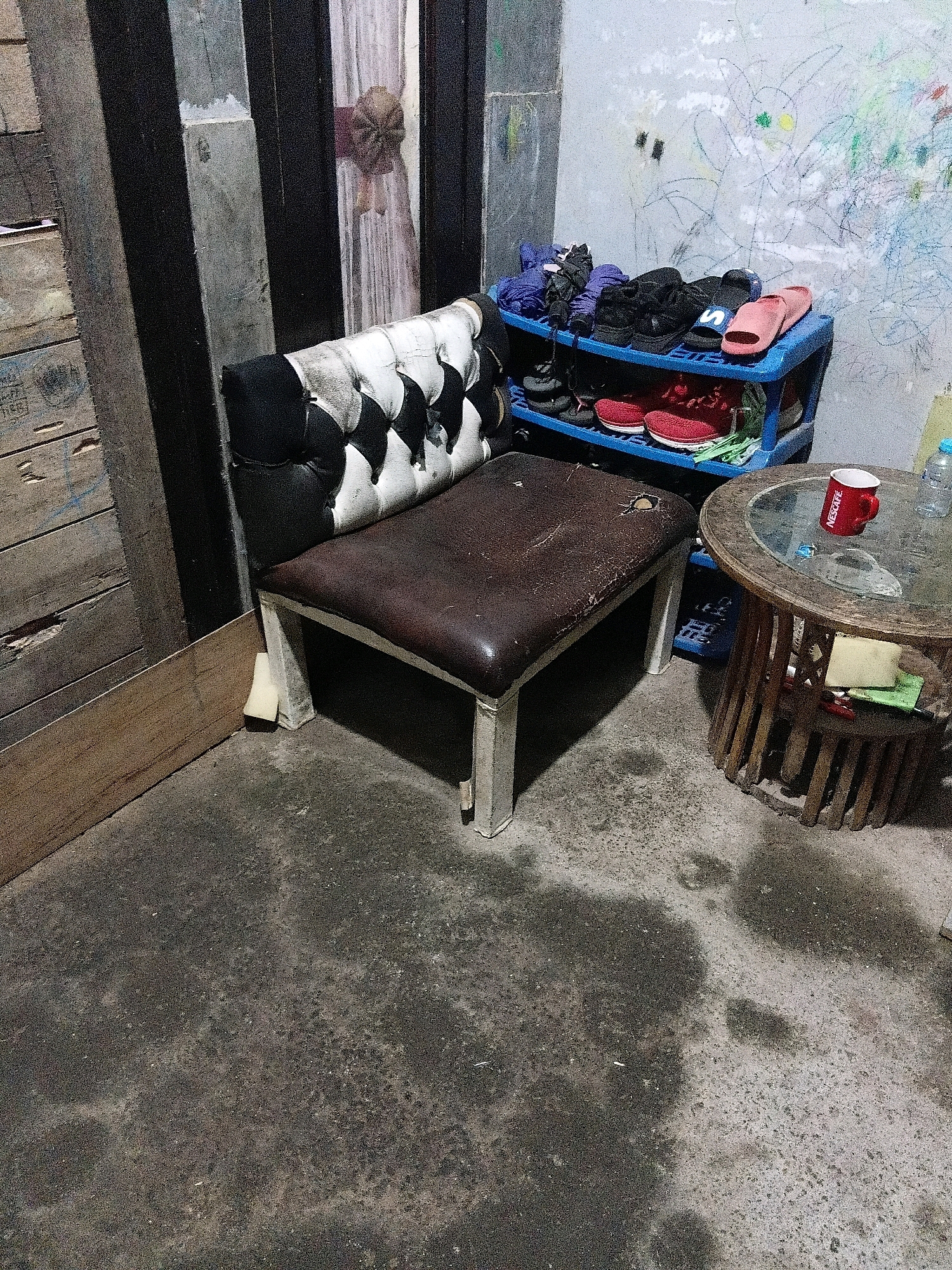 The image depicts a cluttered indoor space, possibly a room or a covered outdoor area, in Cikal, Indonesia. In the foreground, a worn, tufted chair with a brown leather seat and a black and white patterned backrest sits on a rough concrete floor. The chair's legs are visible, with one leg propped up by a piece of foam. To the right of the chair is a round wooden table with a glass top. On the table, there is a red "Nescafe" mug, a bottle of water, and some papers. Behind the chair and table, a blue plastic shoe rack is filled with various shoes and footwear, including pink sandals, red sneakers, and black shoes. The wall behind the shoe rack is covered in colorful, childlike crayon drawings. To the left of the chair, a wooden structure made of planks is visible, leaning against a dark, striped vertical surface. The overall impression is one of a utilitarian and lived-in environment.