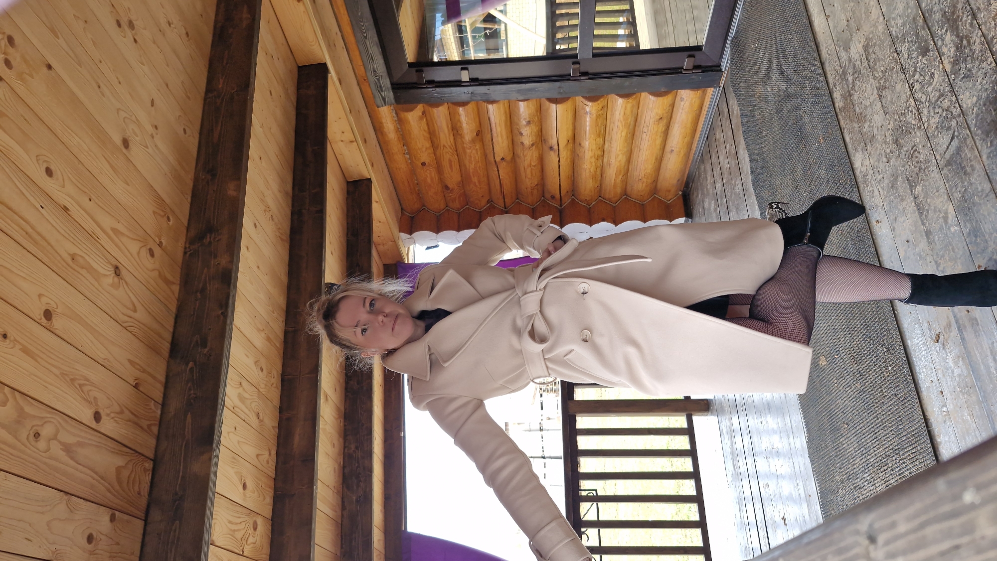 A woman is depicted posing inside a rustic wooden structure, likely a porch or veranda, in Otchor, Russia. She is wearing a long, light beige trench coat, cinched at the waist with a tied belt, over what appears to be a dark top. Her lower body is covered in black fishnet stockings and black ankle boots; one boot features a distinctive patterned heel, possibly snakeskin. Her hands are either in her coat pockets or subtly holding the belt, contributing to a relaxed yet posed stance. She has light-colored hair, styled loosely, and looks directly at the camera with a neutral to slightly thoughtful expression.
The setting is characterized by extensive use of natural wood. The walls and ceiling are made of light-colored wooden planks and horizontal logs, creating a warm, cabin-like aesthetic. Darker wooden beams are visible throughout the structure, adding architectural contrast. The floor consists of wooden planks, with a dark, textured mat near a doorway or large window. Through this opening, a metal railing and an indistinct outdoor view are visible, suggesting the structure is elevated or overlooks an area. The natural lighting indicates it is daytime, with clear weather outside. No visible text is present in the image.