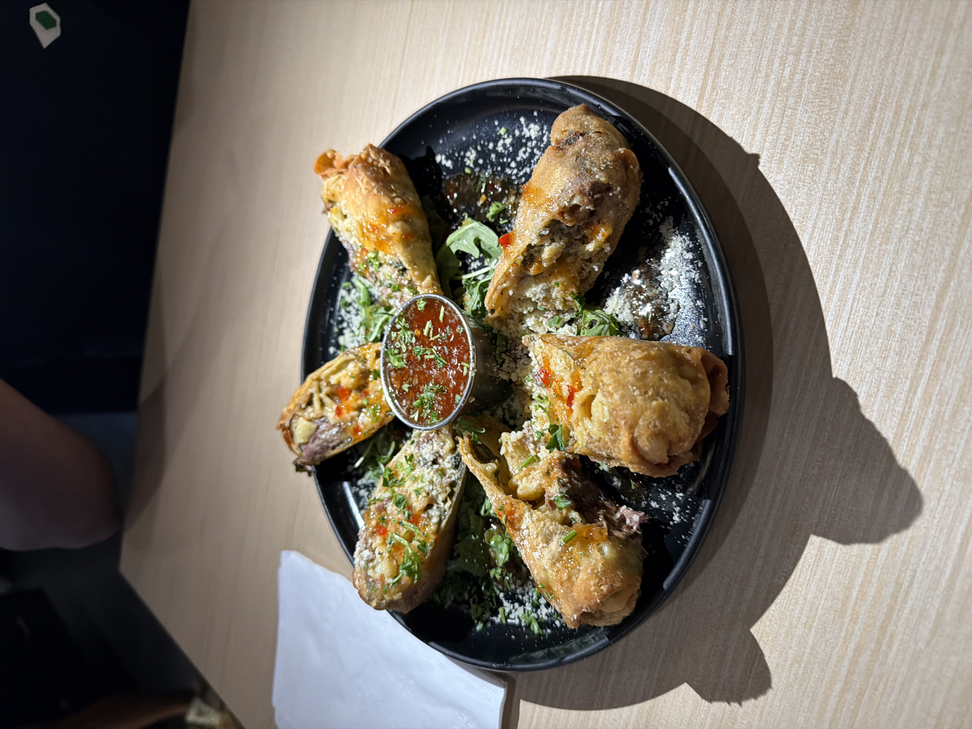 A high-angle shot depicts a dark plate centrally positioned on a light wooden table, likely indoors. The plate holds six food items that appear to be fried dumplings or spring rolls, arranged in a circular fashion. A small, metal container filled with a red sauce is placed at the center of the circle. The food is garnished with herbs and possibly grated cheese. A shadow is cast from the plate onto the table, and parts of two individuals wearing black tops are visible on the left side of the frame. A white napkin sits near the bottom left corner. Without additional information, the exact location within Los Angeles, United States, cannot be determined.