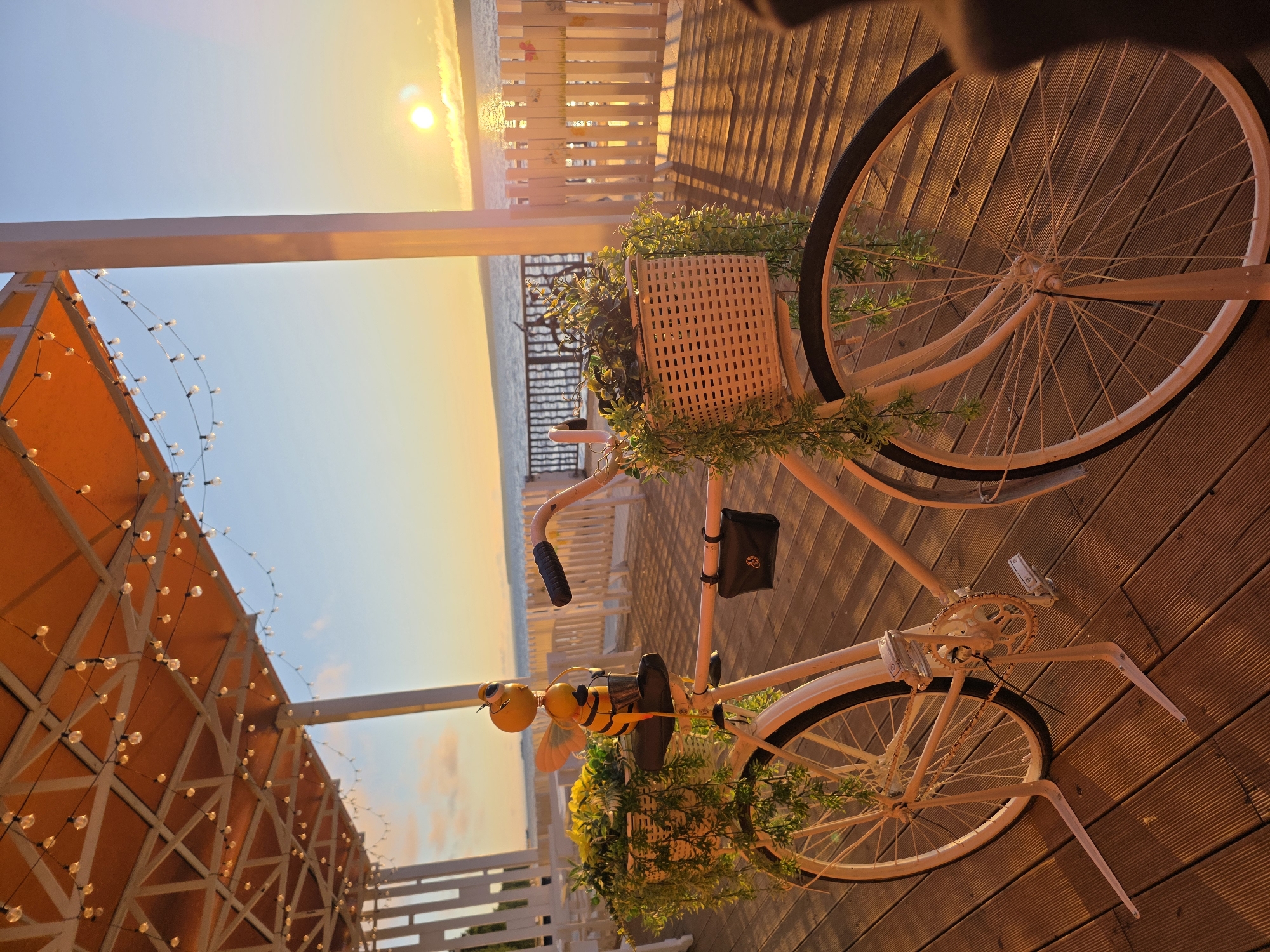 The image captures a decorative bicycle placed on a wooden deck, likely part of an outdoor seating area at a cafe or restaurant. The bicycle is white with a basket in the front filled with yellow flowers and trailing greenery. A decorative bee figurine is perched on the seat. Behind the bicycle, a white picket fence separates the deck from a body of water, possibly a river or lake, with a sunset visible in the background. The scene is illuminated by warm, golden hour light. The location is identified as Chaikovsky, Russia, suggesting this is an outdoor establishment within the city. There are no people or actions depicted in the image.