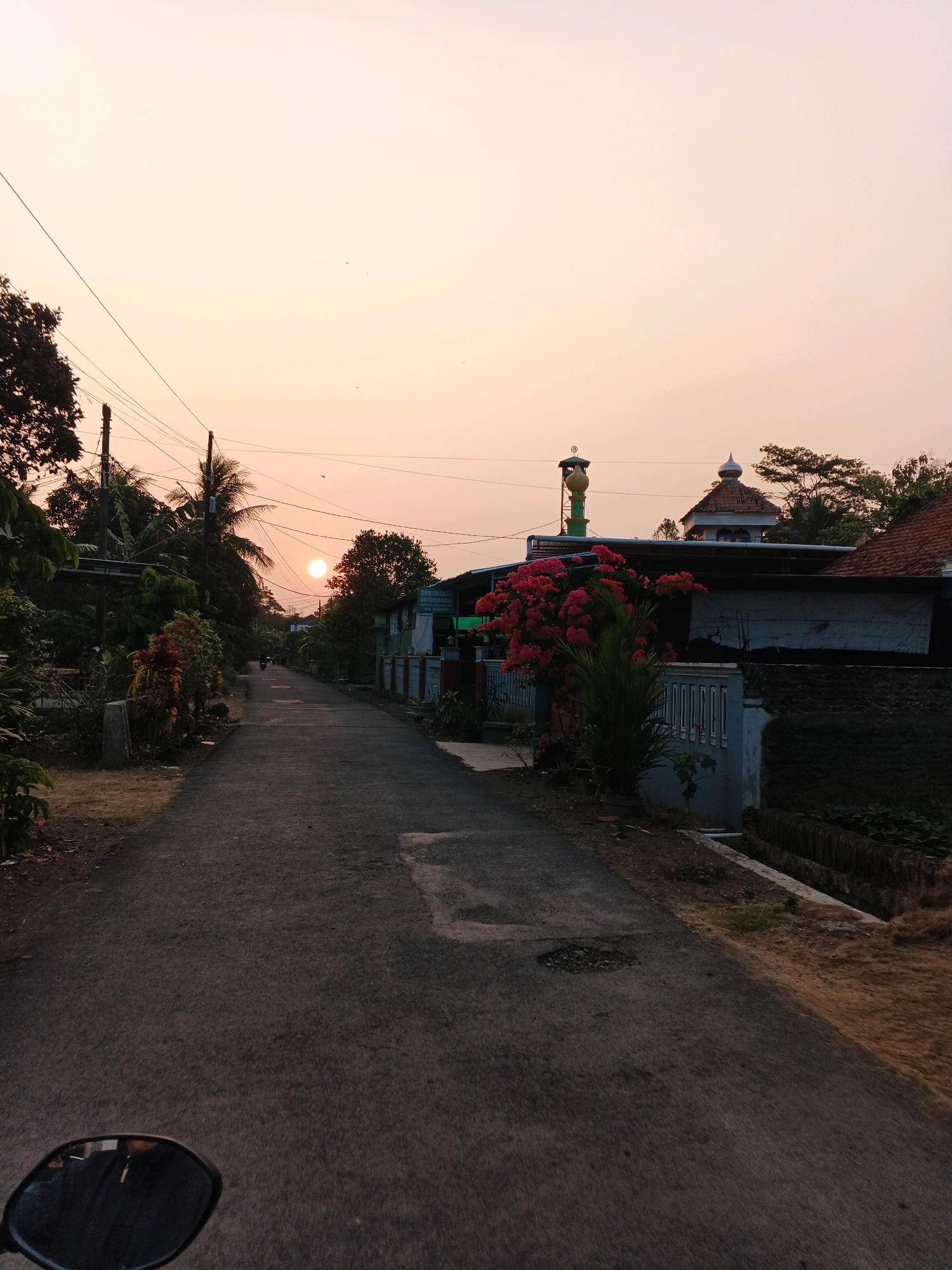 The image depicts a rural road at sunset in Kesugihan, Indonesia. The sun, a bright orb, is low in the sky, casting a warm glow. A paved road stretches into the distance, lined with houses and lush vegetation. A mosque with a green minaret and a domed roof is visible in the background. The foreground shows a motorcycle's rearview mirror, suggesting the photo was taken from a moving vehicle. The scene is peaceful and serene, capturing the quiet beauty of village life.