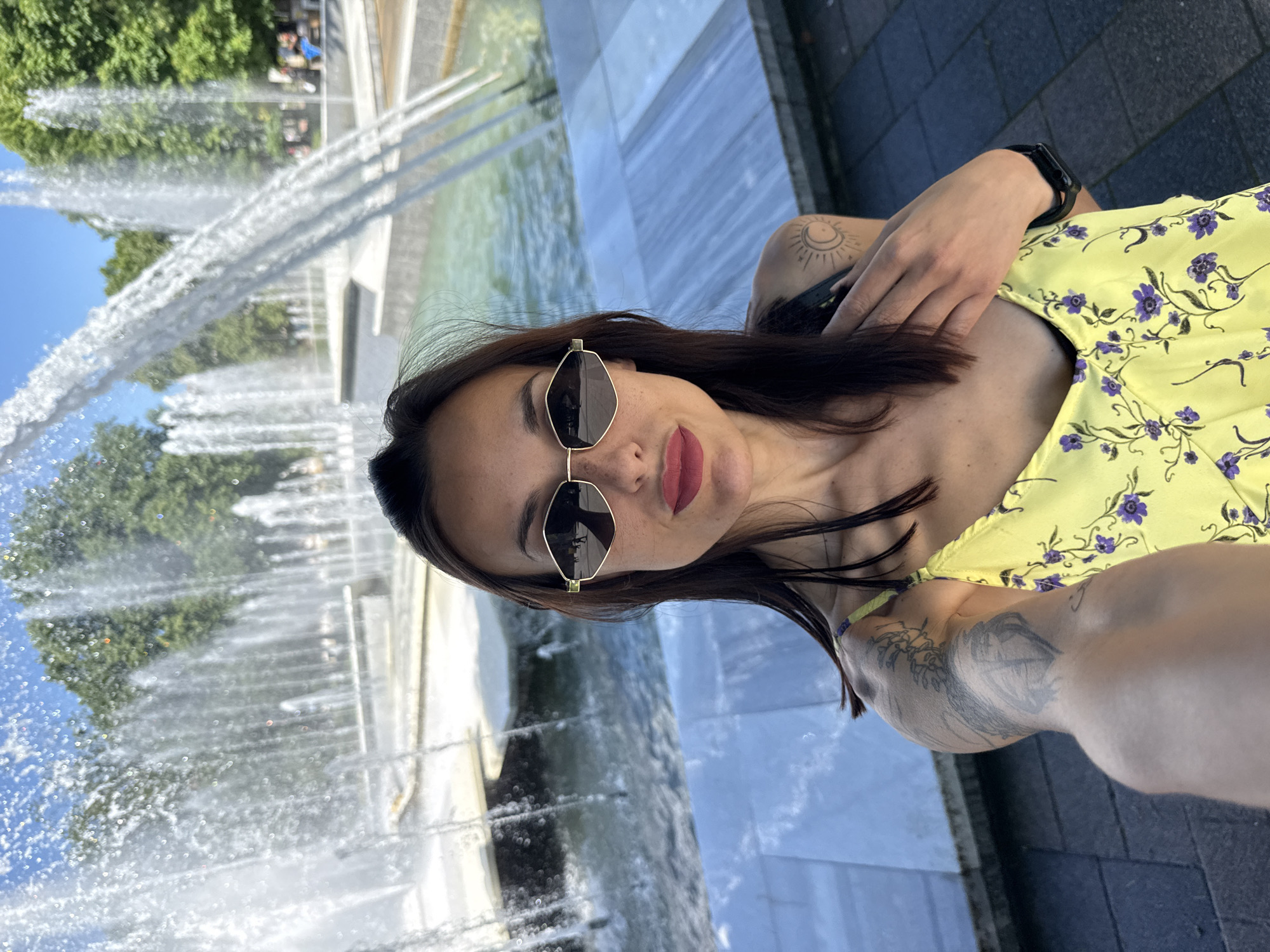 A woman in a yellow floral dress and sunglasses stands in front of a large fountain in Pleven, Bulgaria, taking a selfie. The fountain features multiple jets of water, and trees are visible in the background. The woman has brown hair and tattoos on her arms. She is wearing a smartwatch on her left wrist. The ground is paved with gray tiles.