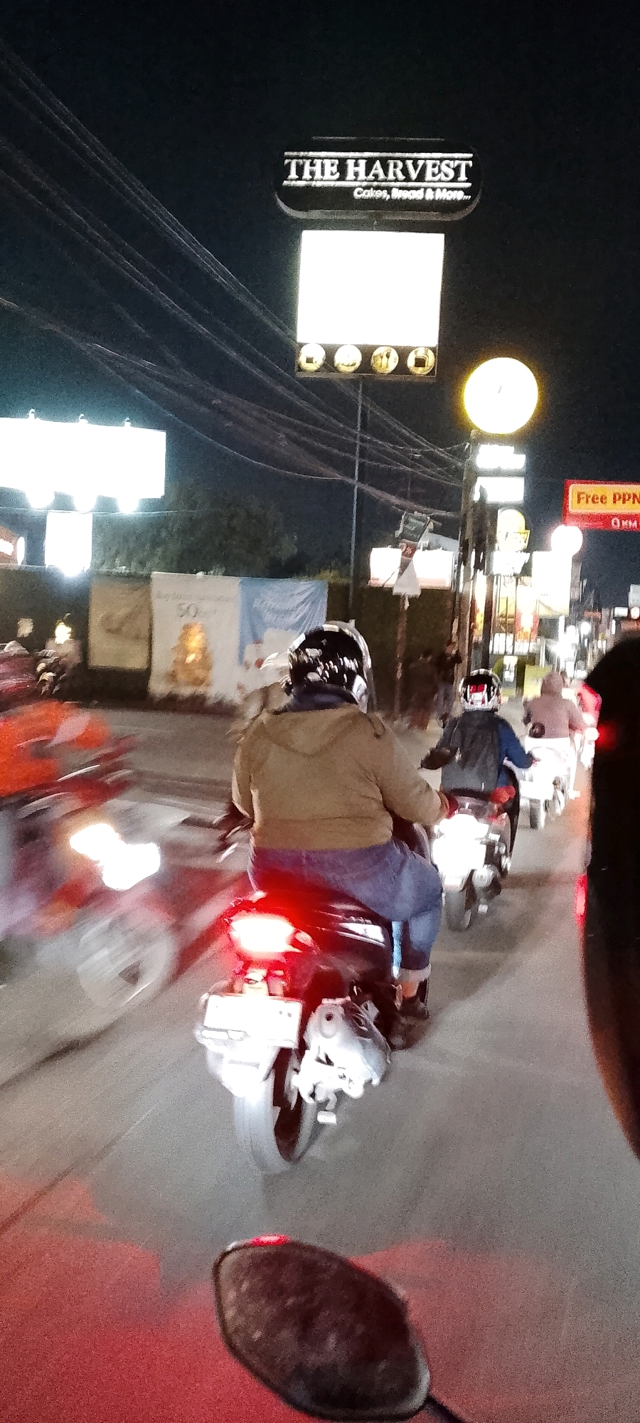 The image shows a nighttime street scene in Gunung Putri, Indonesia. Several motorcycles are in motion on a road with streetlights and illuminated signs. In the foreground, a person wearing a brown hoodie and blue jeans rides a motorcycle, their back to the camera. The motorcycle's brake light is illuminated red. To the right and slightly behind, another person on a motorcycle is visible, wearing a dark jacket and helmet. Further ahead, other motorcycles and indistinct figures can be seen. A prominent sign above the road reads "THE HARVEST" with smaller text below indicating "Cakes, Bread & More...". Other illuminated signs and billboards are scattered throughout the scene. The environment suggests an urban or semi-urban commercial area at night.