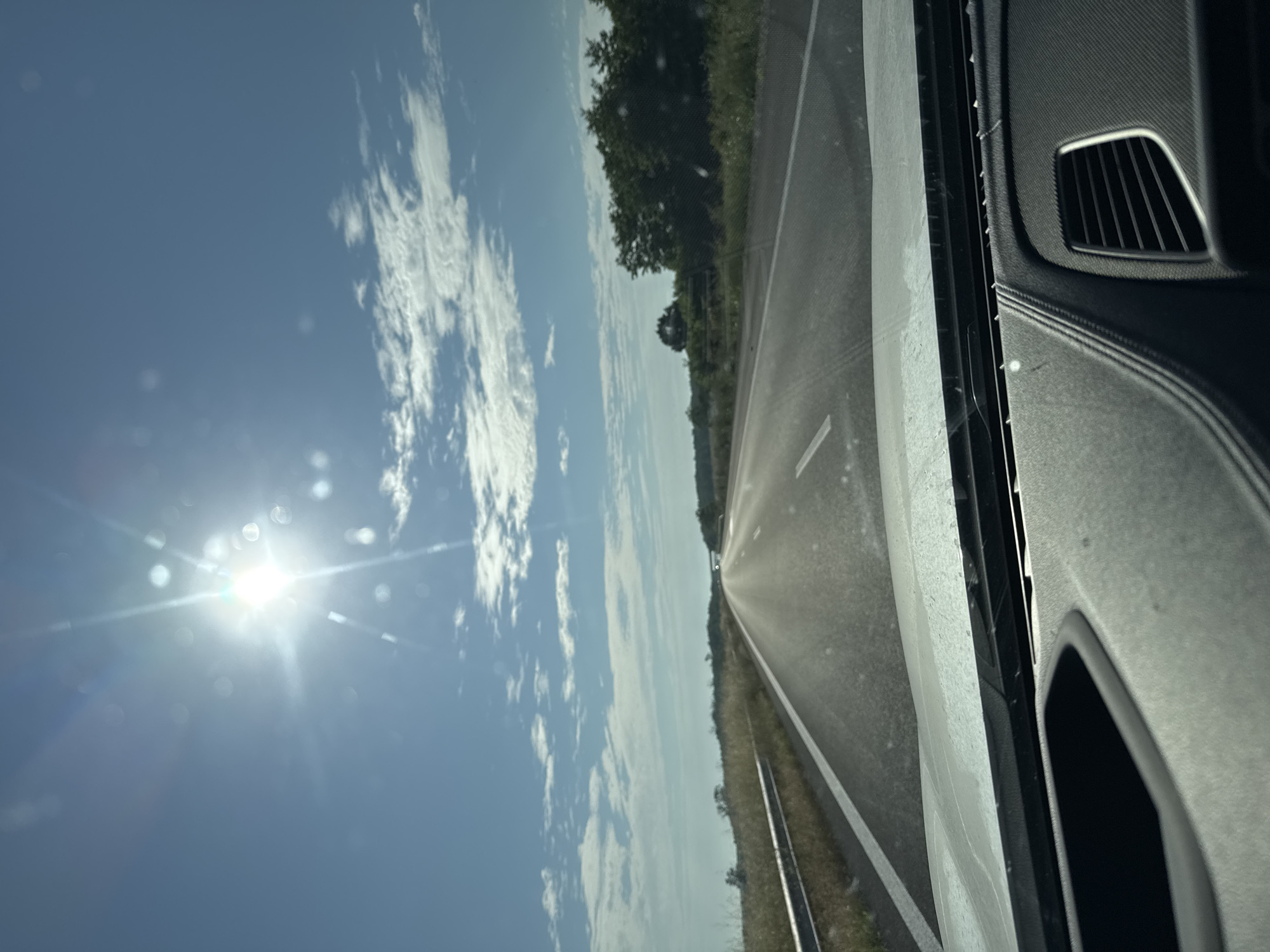 The image displays a forward-facing view from inside a vehicle, looking out onto a highway under bright daylight. The sky is predominantly clear blue with scattered white cumulus clouds, primarily in the upper right quadrant. The sun is intensely bright in the upper-left-center, causing significant lens flare and glare that diffuses light across the upper frame.
The asphalt highway appears straight and extends into the distant horizon, with a noticeable shimmering effect on its surface. White solid lines mark the road's shoulder and potentially separate lanes. To the left, a silver metal guardrail runs parallel to the road, beyond which lies dry, yellowish-brown grass and low vegetation. The right side of the road also features dry grasses, leading to a dense line of green trees in the mid-distance, defining the horizon on that side. No people are visible.
The bottom portion of the image shows the dark gray dashboard of the vehicle. It features distinct textures, an open compartment or speaker grille on the left, and a multi-slatted air vent on the right, consistent with a contemporary car interior. The windshield exhibits minor specks or smudges.
This scene depicts a rural or semi-rural highway environment, characterized by open land, trees, and clear weather. Based on the provided instruction, this highway setting is located within or in the vicinity of Kolarovo, Bulgaria.