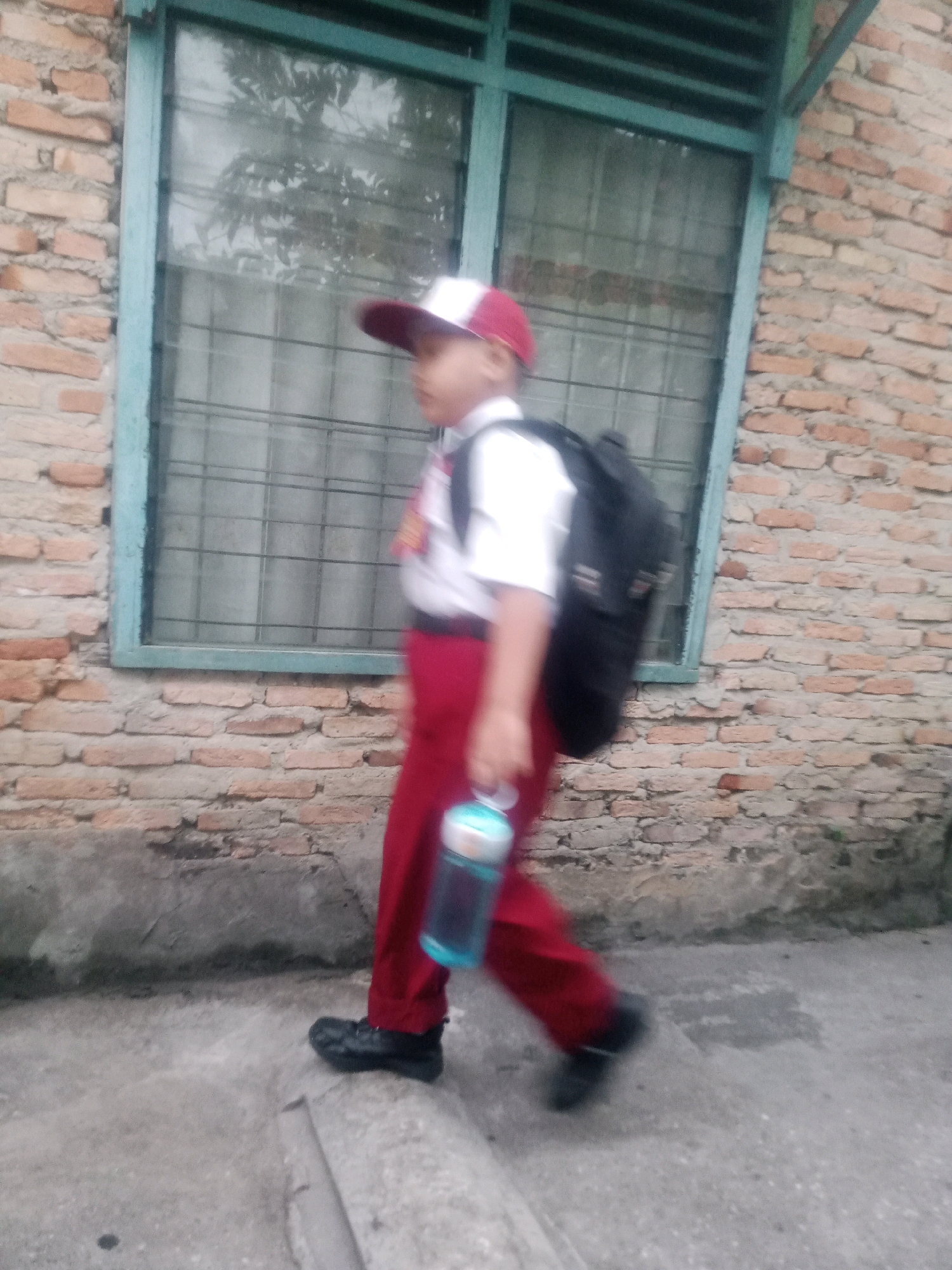 A blurred, medium shot captures a child walking with a backpack near a brick wall and a window. The child, wearing a white shirt, red pants, a red and white hat, and black shoes, carries a light blue water bottle in their right hand. The background consists of a rough brick wall and a window with a green frame and metal bars. The ground is paved with concrete. The scene is consistent with an outdoor setting in Pekanbaru, Indonesia, possibly near a school or residential building. The image quality is poor due to motion blur.